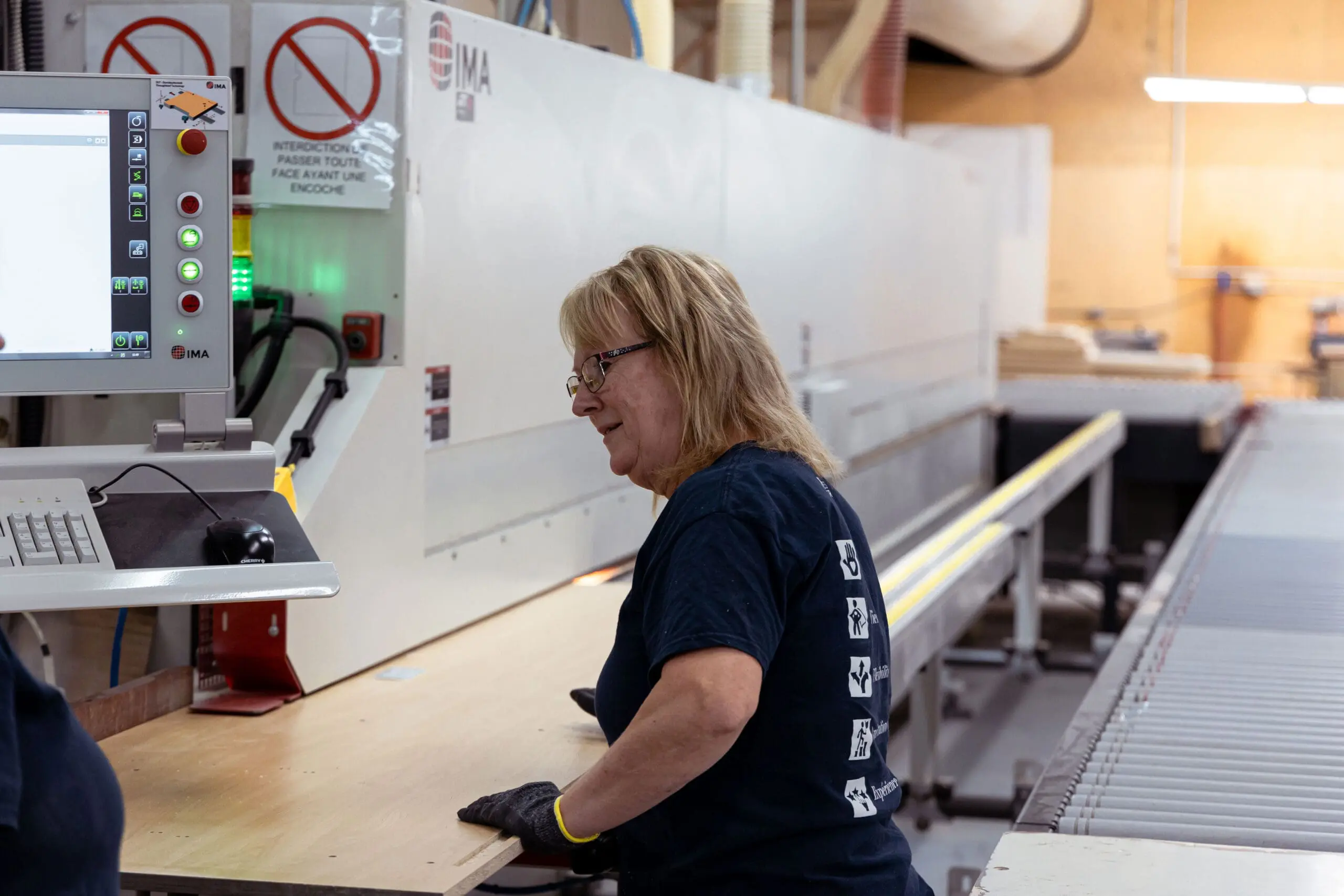 Une femme portant des gants et des lunettes de sécurité fait fonctionner des machines dans une usine, introduisant une planche de bois dans une grande machine industrielle ; un écran d'ordinateur et des panneaux de sécurité sont visibles à proximité.