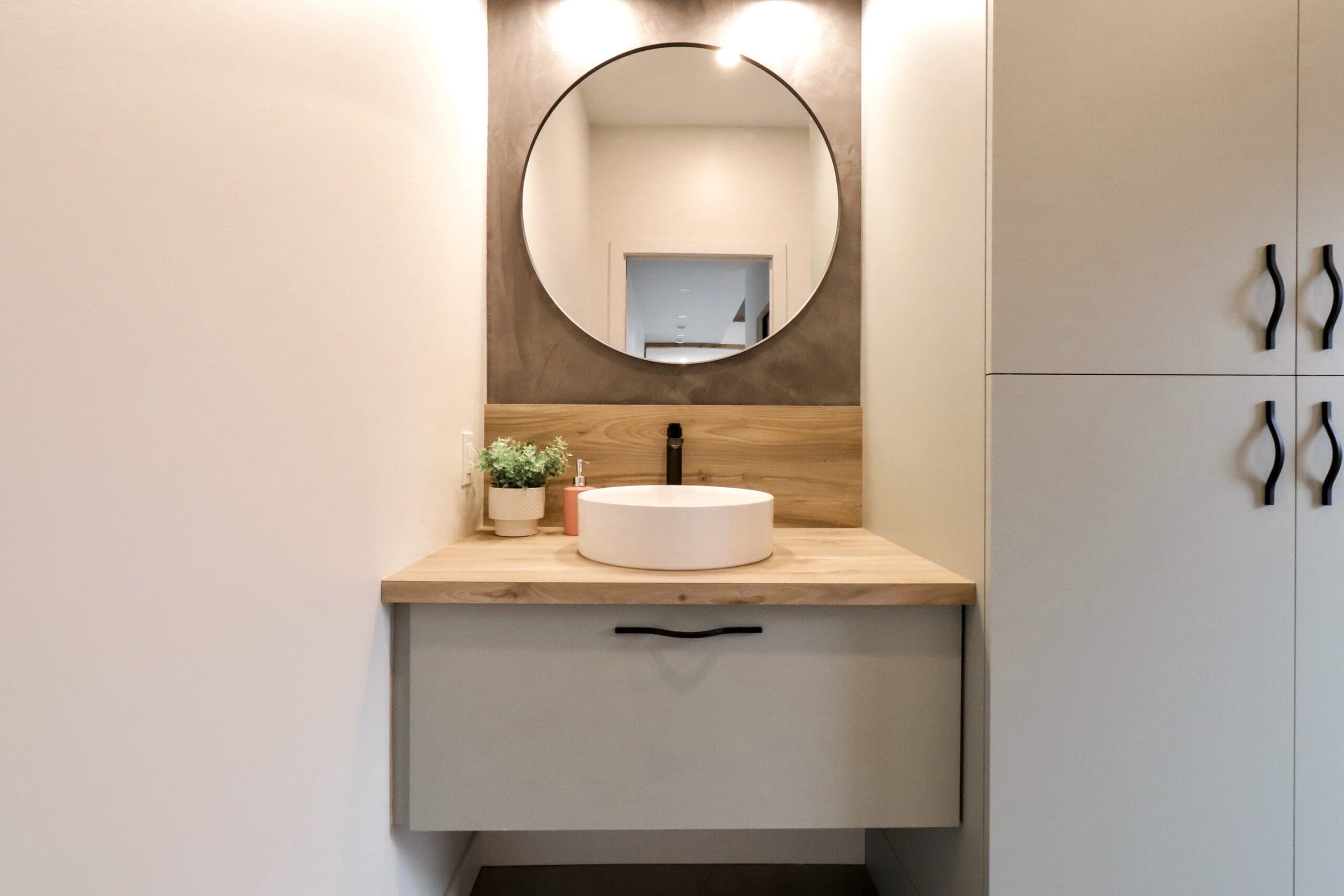 Meuble de salle de bain moderne avec miroir rond, comptoir en bois, évier blanc, robinet noir et plante en pot. Une lumière éclaire le miroir et une armoire avec des poignées noires se trouve à droite.