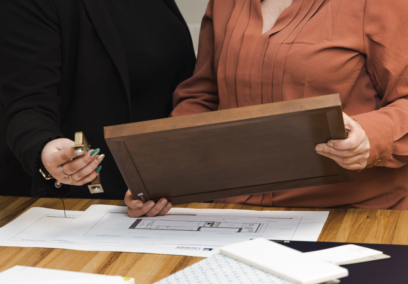 Deux femmes sont assises à une table et examinent des plans de conception et un échantillon de porte d'armoire en bois. L'une d'elles montre les plans tandis que l'autre tient l'échantillon, toutes deux souriant dans un espace de travail moderne et lumineux.