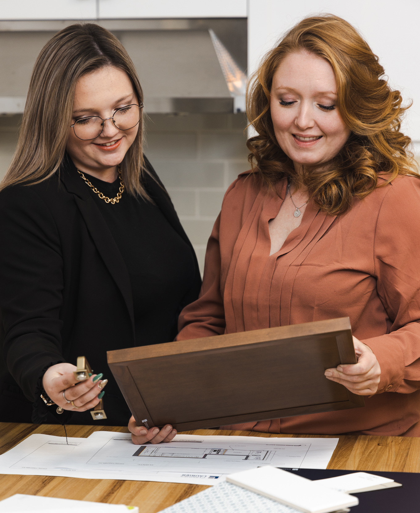 Deux femmes sont assises à une table et examinent des plans de conception et un échantillon de porte d'armoire en bois. L'une d'elles montre les plans tandis que l'autre tient l'échantillon, toutes deux souriant dans un espace de travail moderne et lumineux.