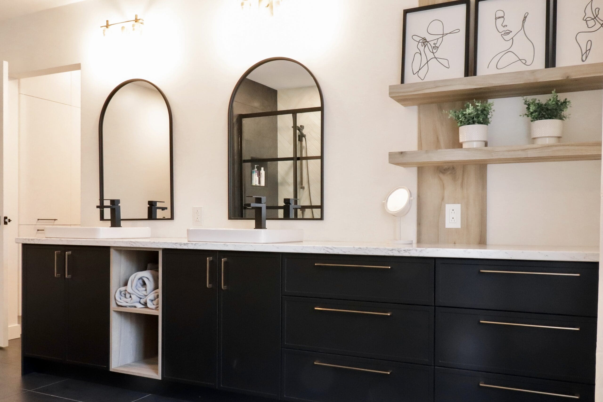 Salle de bains moderne avec double lavabo, armoires noires avec poignées dorées, deux miroirs cintrés, art minimaliste au mur, étagères avec plantes en pot, et serviettes soigneusement enroulées dans une étagère ouverte.