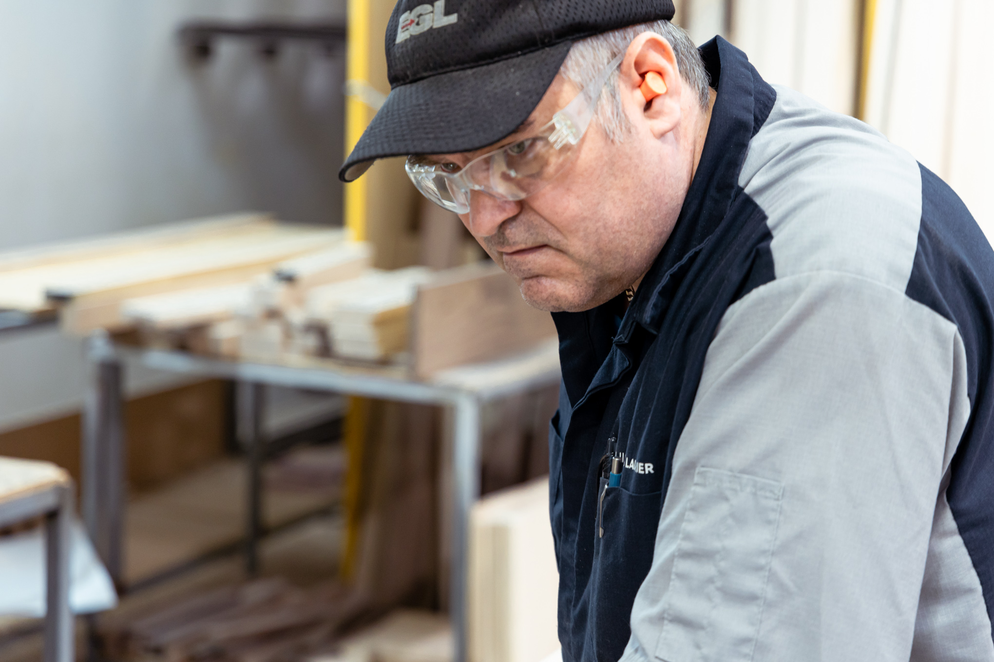Un homme portant des lunettes de sécurité, des protections auditives et une casquette noire travaille intensément dans un atelier de menuiserie avec des pièces de bois et des outils en arrière-plan.
