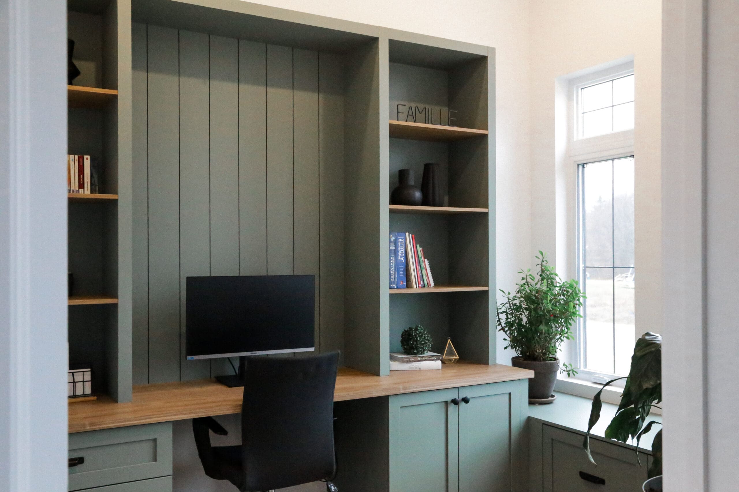 Un bureau à domicile moderne avec des étagères et des armoires encastrées vert sauge, un bureau en bois, un écran d'ordinateur, des livres, des objets décoratifs et une plante en pot près d'une grande fenêtre laissant entrer la lumière naturelle.