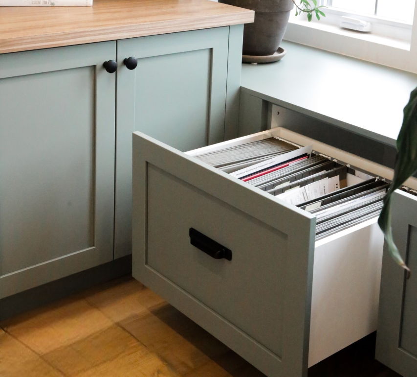 Le tiroir d'un classeur vert est ouvert, laissant apparaître des dossiers bien rangés. L'armoire se trouve à côté d'une fenêtre avec une plante en pot sur le rebord et une étagère avec quelques livres et objets décoratifs sur le dessus.
