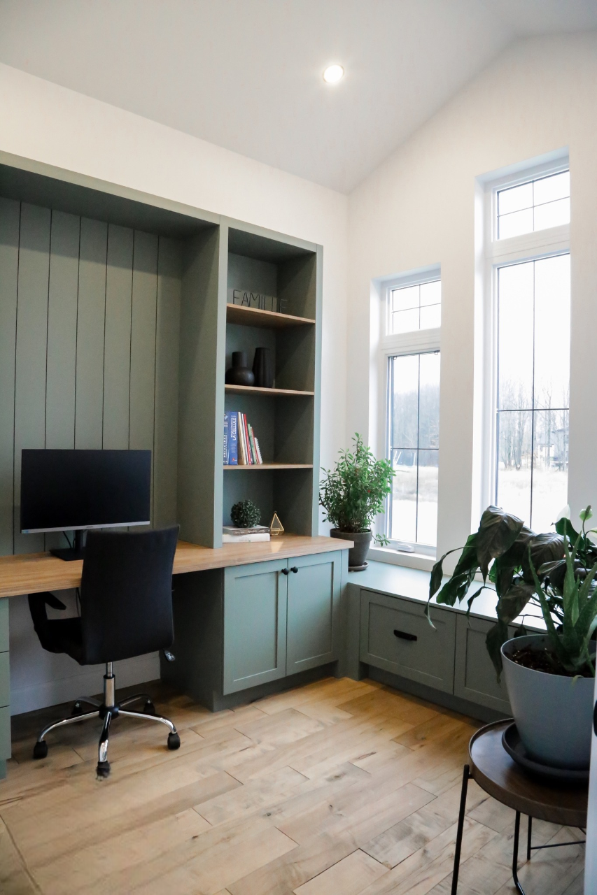 Un bureau à domicile moderne avec des étagères vertes encastrées, un écran d'ordinateur sur un bureau en bois, une chaise noire, des plantes en pot, des livres et de grandes fenêtres laissant entrer la lumière naturelle.