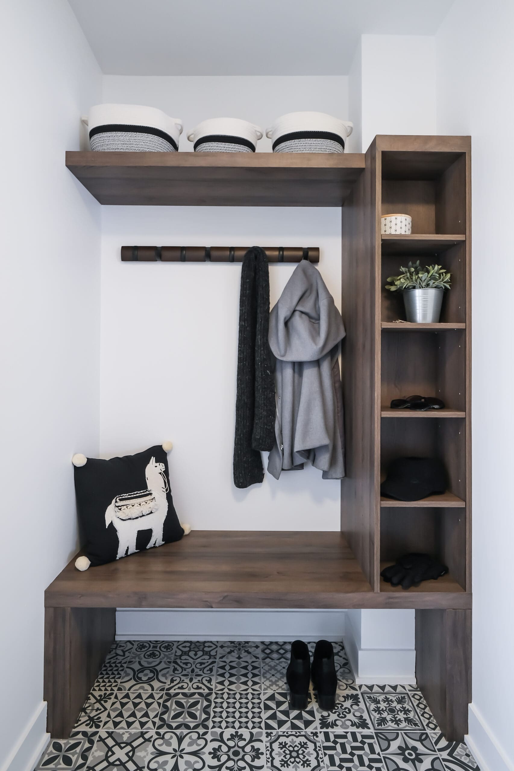 Un vestiaire moderne avec un sol en carrelage à motifs, un banc en bois, des étagères, des paniers de rangement, une plante, des patères avec des vestes, un coussin avec un motif de lama et une paire de chaussures noires sur le sol.