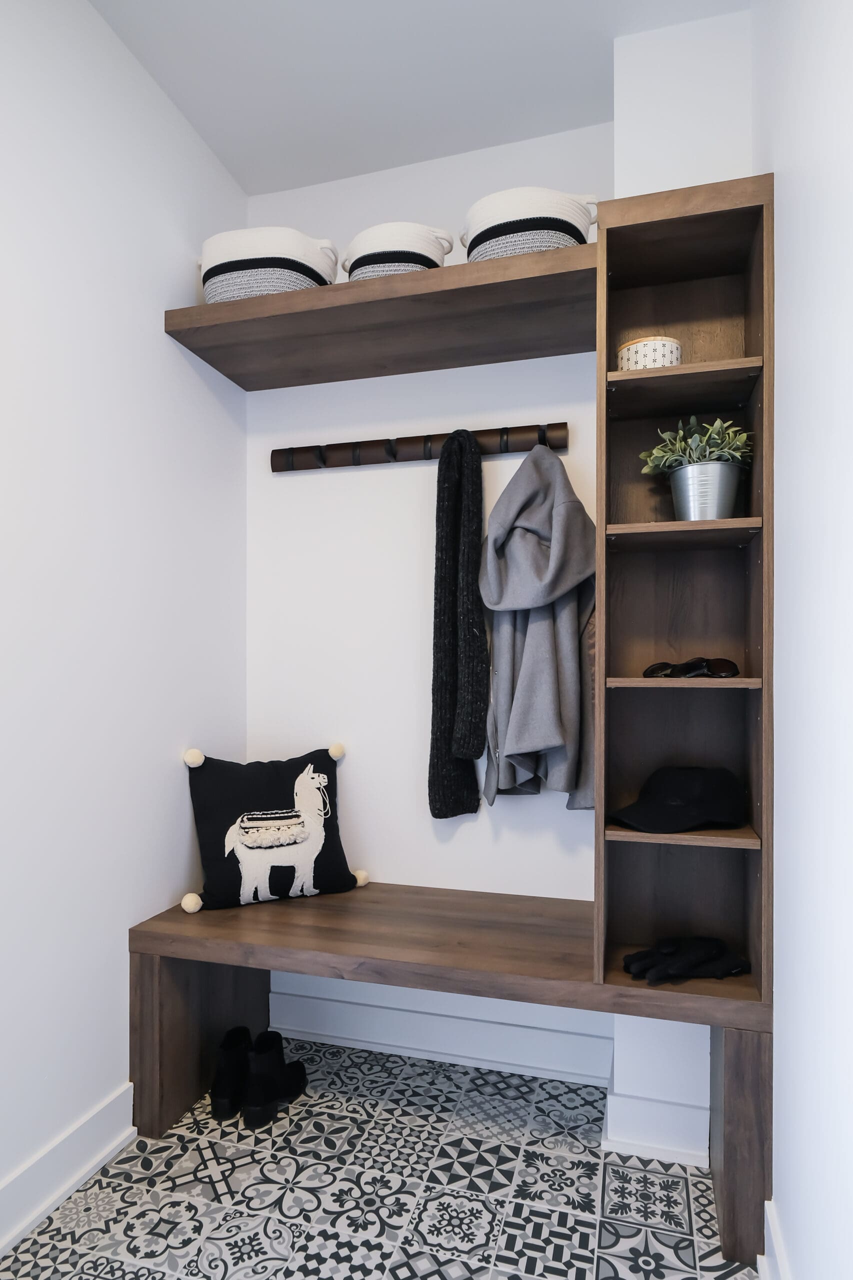 Une petite entrée présente un carrelage à motifs, un banc en bois avec un oreiller en forme de lama, des étagères ouvertes avec des paniers, des patères avec des vestes, une plante en pot, des chapeaux et des chaussures bien rangées.
