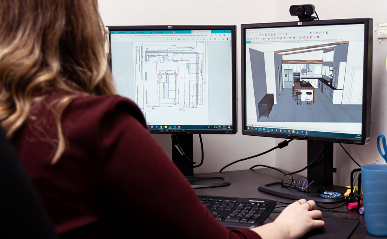 Une personne travaille à un bureau équipé de deux écrans d'ordinateur. L'un des écrans affiche des plans architecturaux, tandis que l'autre montre un rendu en 3D de la décoration intérieure d'une cuisine moderne.