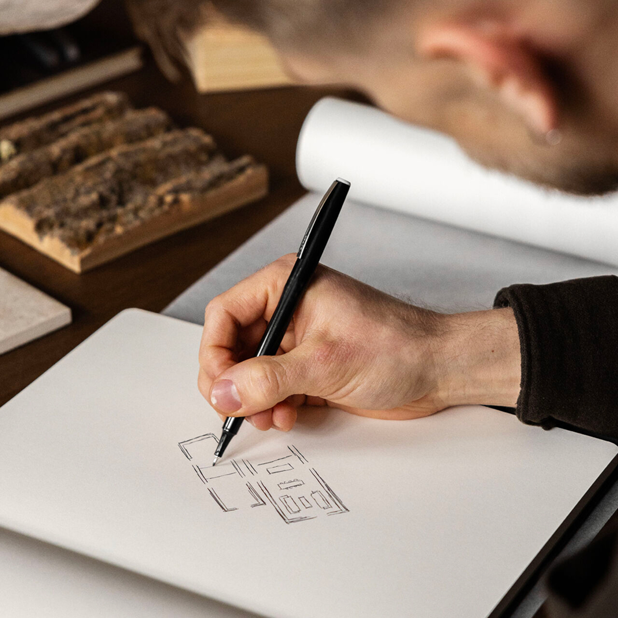 Une personne esquisse un dessin géométrique au stylo noir sur un carnet blanc vierge, avec des échantillons de bois et d'autres objets flous en arrière-plan.