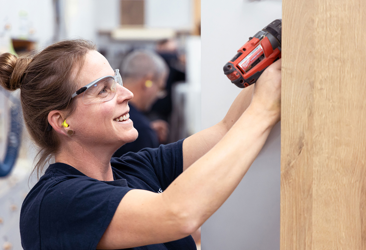 Une femme portant des lunettes de sécurité et des bouchons d'oreille sourit en utilisant une perceuse électrique rouge pour travailler sur un panneau de bois à l'intérieur.