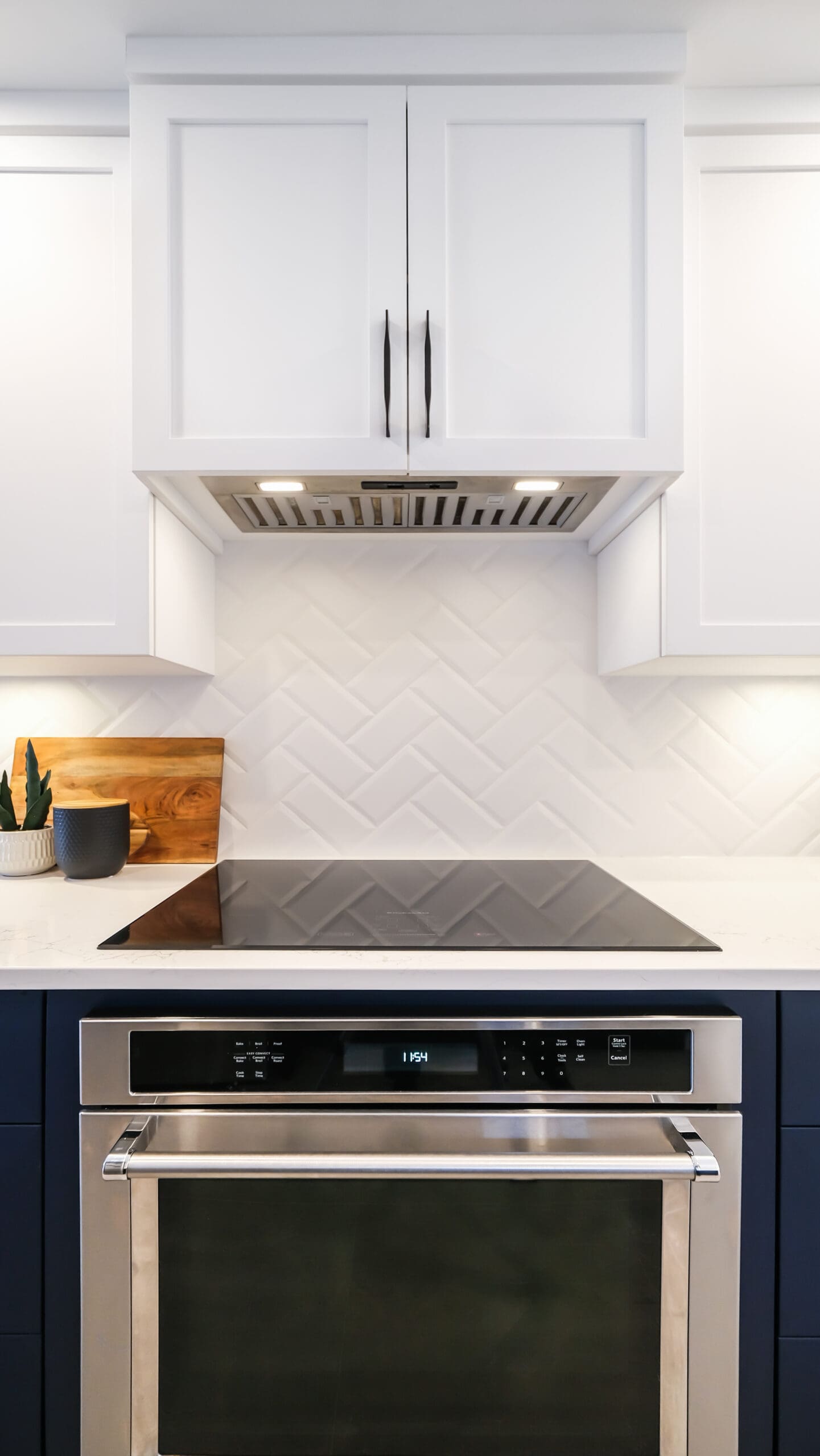 Cuisine moderne avec un four en acier inoxydable, une table de cuisson en verre noir, des armoires blanches et un dosseret en carreaux blancs à chevrons ; une petite plante et une planche à découper se trouvent sur le comptoir.