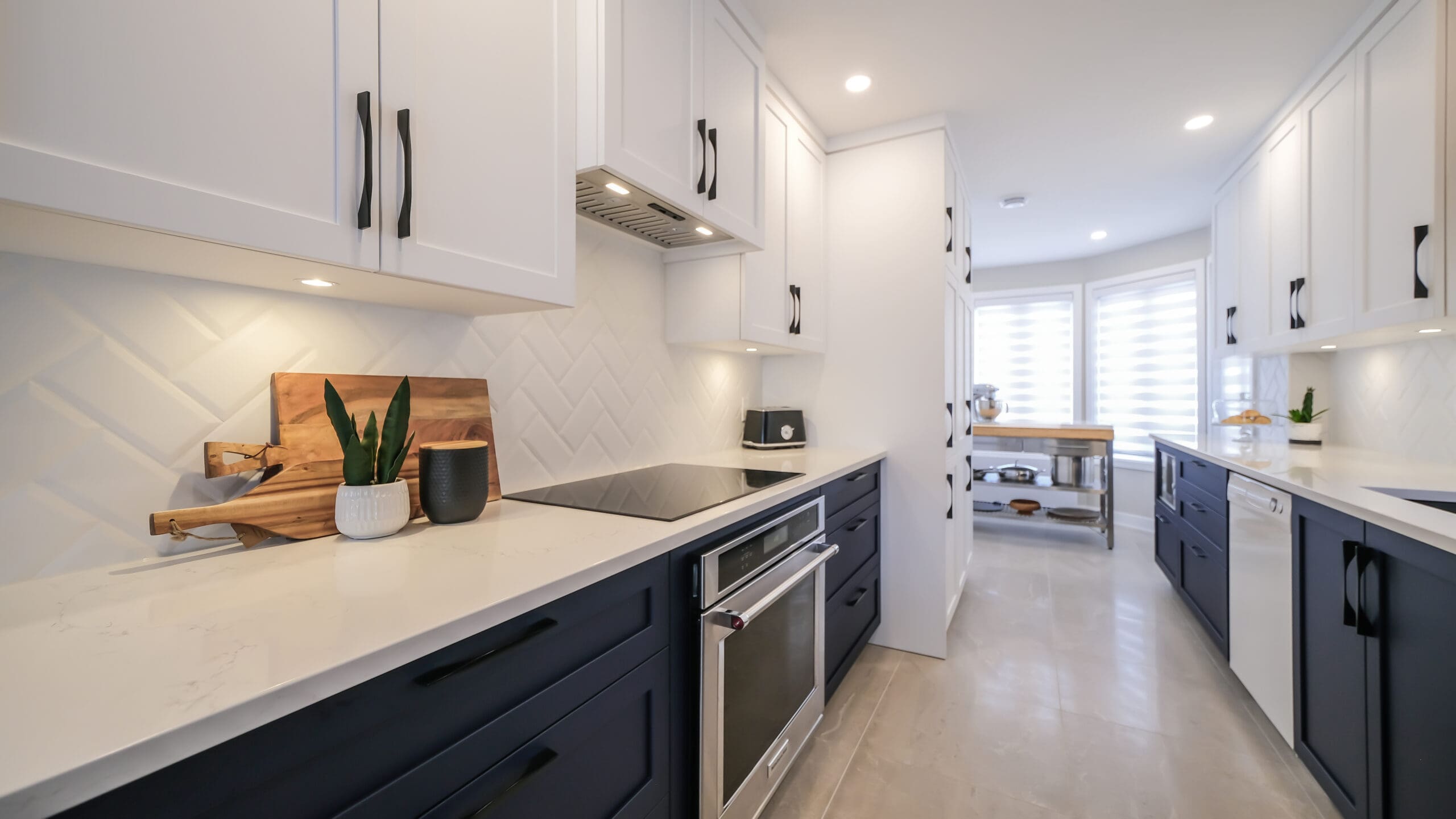 Cuisine moderne avec armoires supérieures blanches, armoires inférieures foncées, comptoirs blancs, cuisinière encastrée, éclairage sous l'armoire et objets décoratifs, menant à un coin repas lumineux avec de grandes fenêtres.
