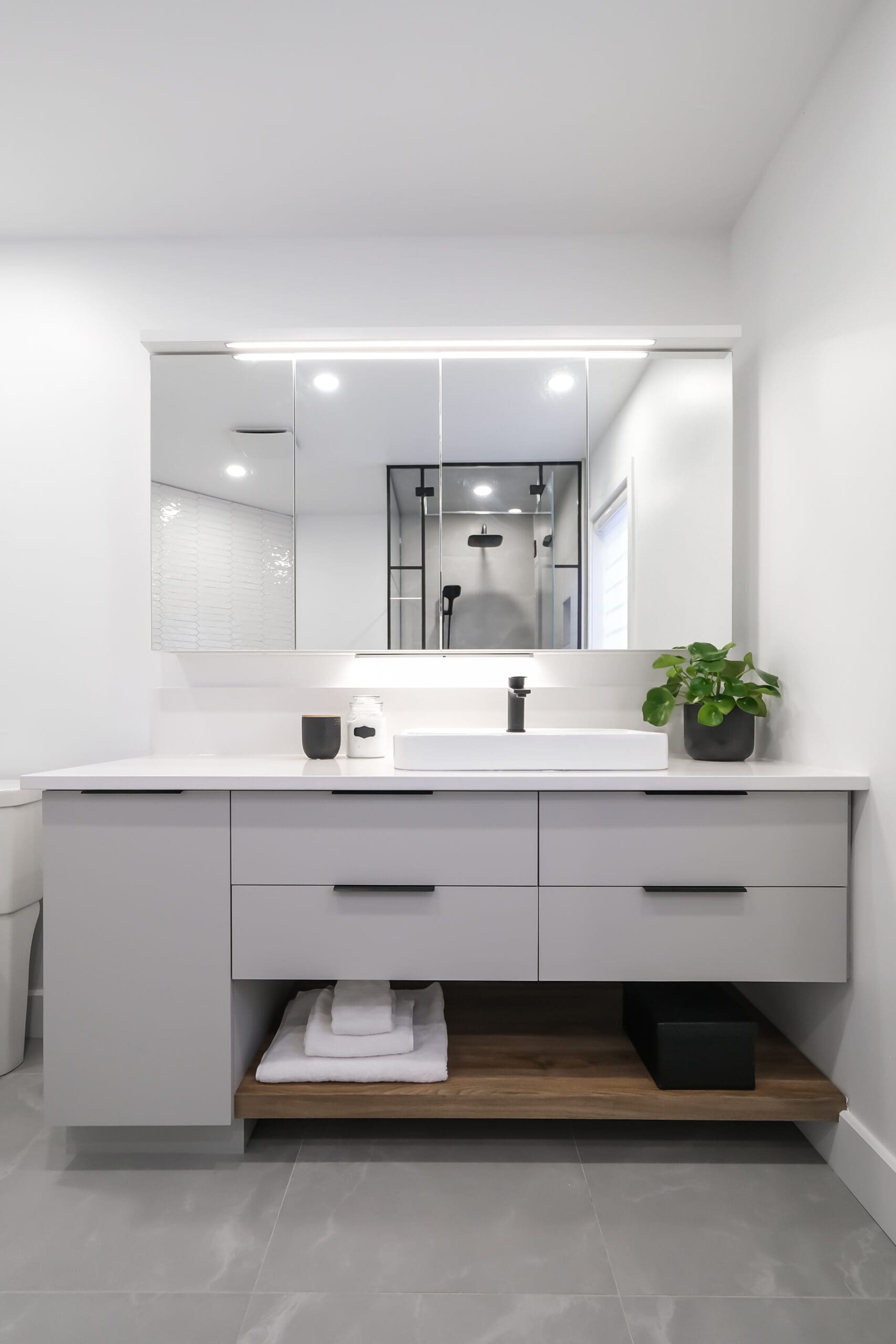 Salle de bains moderne avec un grand miroir, un meuble-lavabo gris, un comptoir blanc, un lavabo, un robinet noir, une petite plante, des serviettes soigneusement pliées sur une étagère en bois et une cabine de douche en verre qui se reflète dans le miroir.