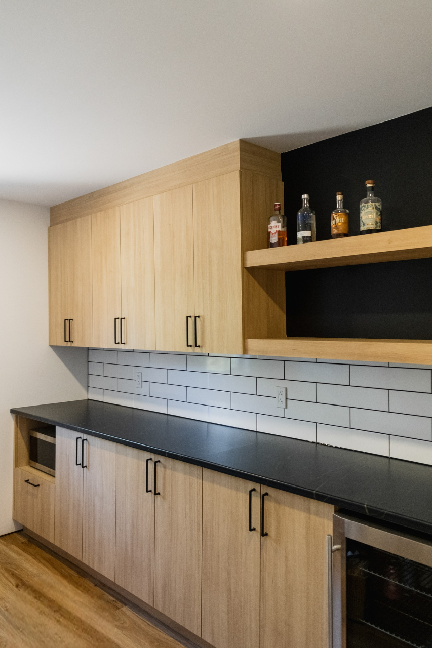 Cuisine moderne avec des armoires en bois clair, un comptoir noir, un dosseret en carreaux de métro blancs, des étagères ouvertes contenant des bouteilles d'alcool et un réfrigérateur à vin encastré sur la droite. Le sol est en bois et les murs sont blancs.
