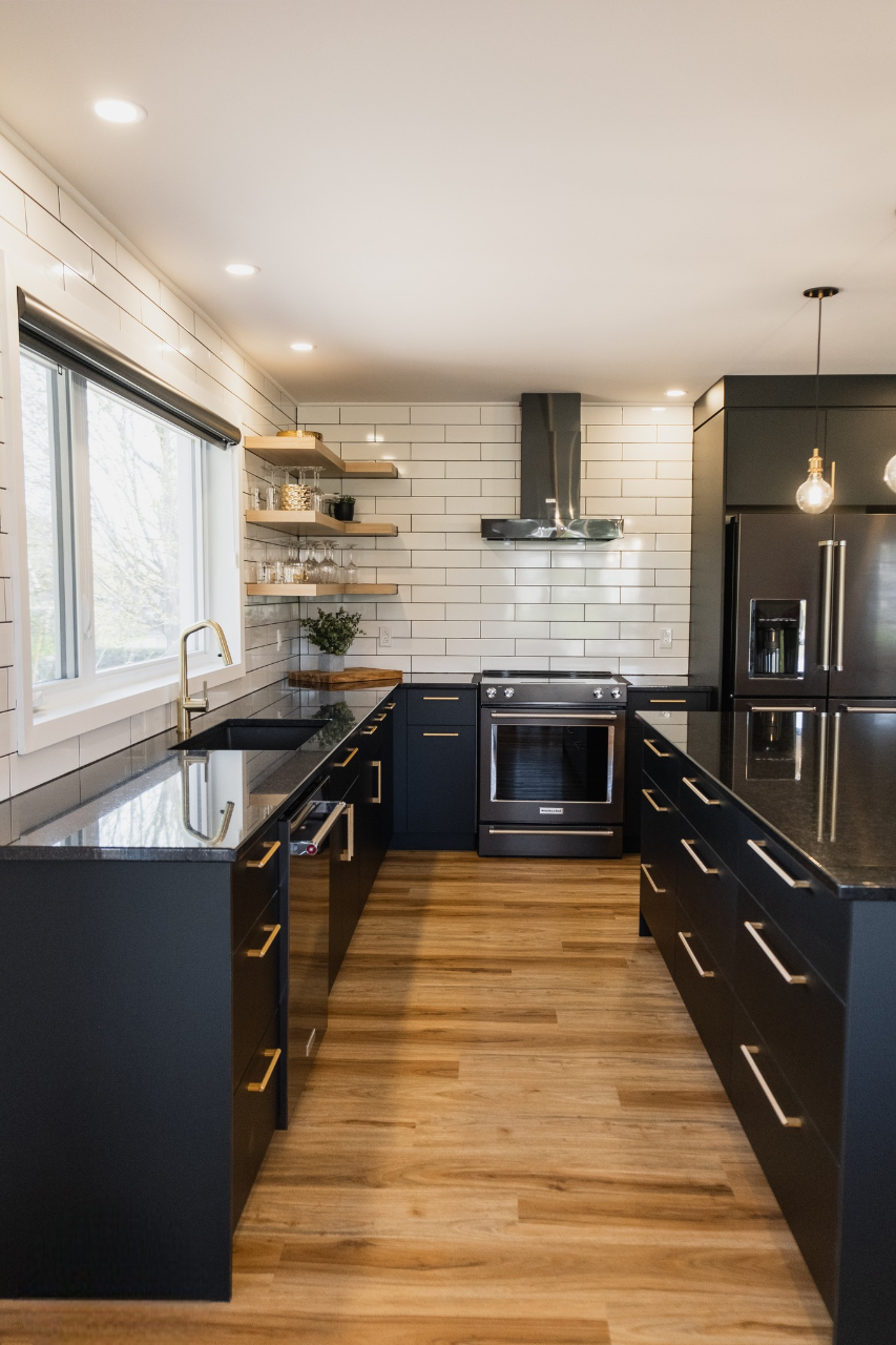 Cuisine moderne avec armoires noires, poignées dorées, appareils électroménagers en acier inoxydable, comptoirs noirs, plancher en bois clair, étagères ouvertes avec vaisselle et dosseret en carreaux de métro blancs. Une grande fenêtre et des lampes suspendues éclairent l'espace.