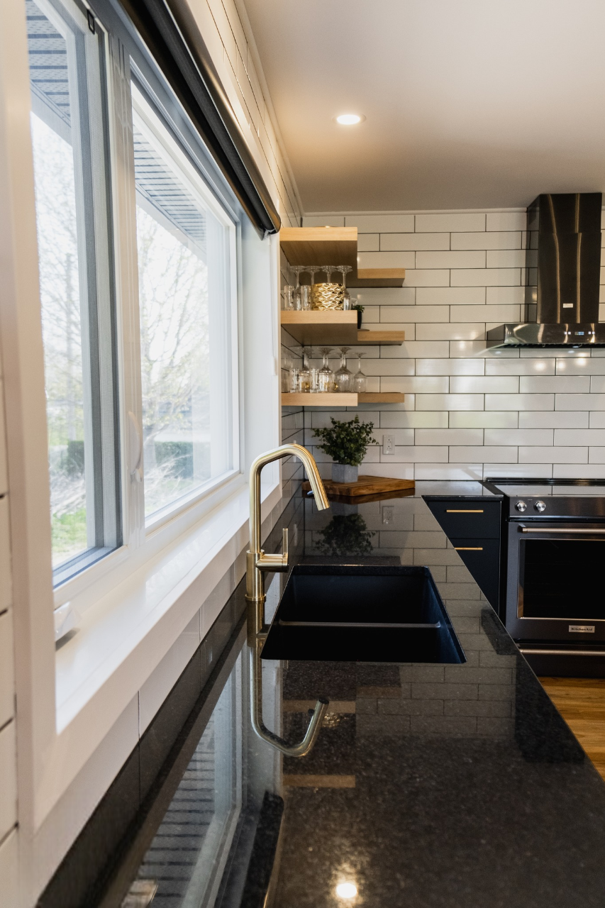Cuisine moderne avec des comptoirs en granit noir, un évier encastré avec un robinet doré, des appareils en acier inoxydable, des étagères ouvertes en bois, un dosseret en carreaux de faïence blancs et une grande fenêtre qui laisse entrer la lumière naturelle.