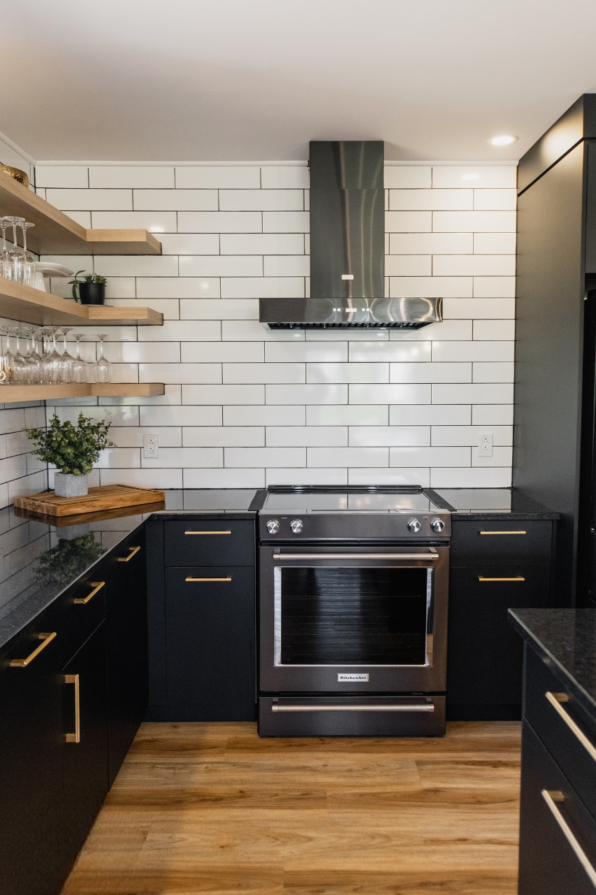 Cuisine moderne avec des armoires noires, des poignées dorées, des étagères en bois pour les verres et les plantes, un dosseret en carreaux de faïence blancs, un four et une hotte en acier inoxydable et un plancher en bois clair.