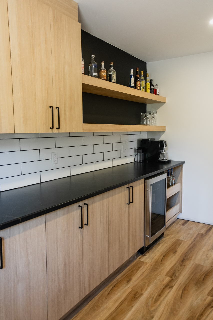Cuisine moderne avec des armoires en bois clair, un comptoir noir, un dosseret en carreaux de métro blancs, des étagères pour les bouteilles et les verres, une cafetière, un mini-réfrigérateur et un plancher en bois.
