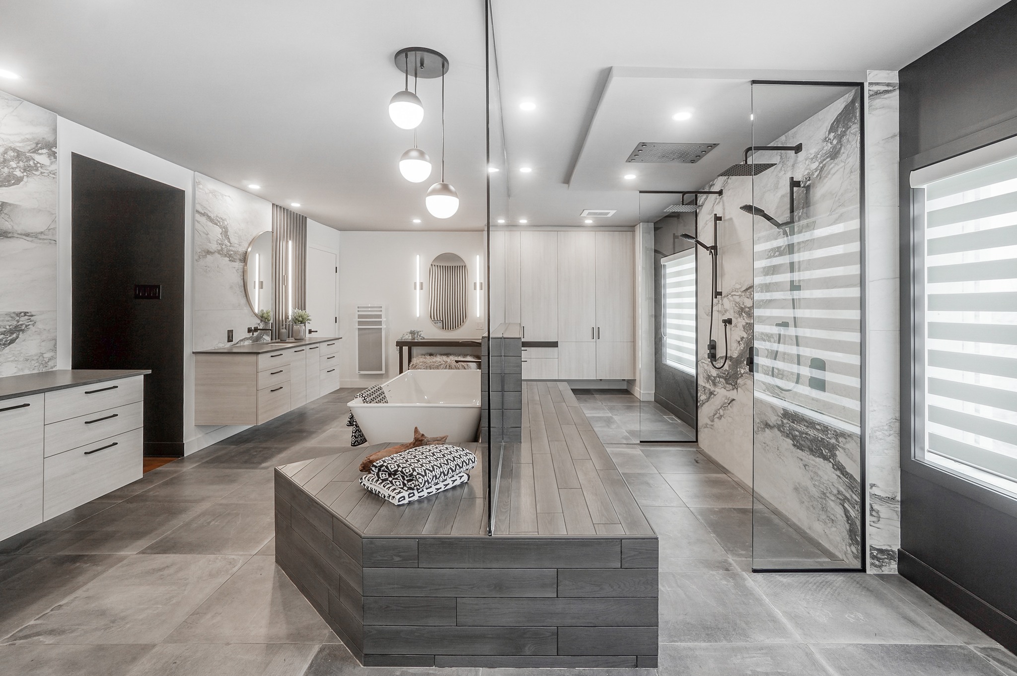 Salle de bains moderne et spacieuse avec douche en verre, baignoire autoportante, double lavabo, grands miroirs, sol en carrelage gris, touches de bois et murs en marbre. Des stores verticaux rayés couvrent une fenêtre sur la droite.