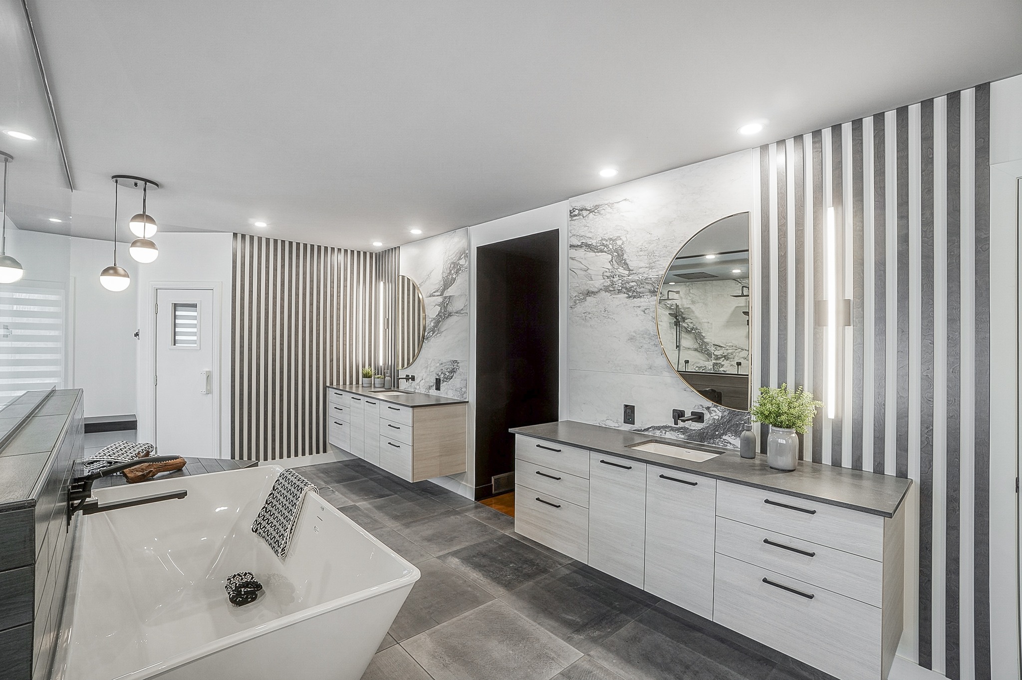 Salle de bains moderne avec une grande baignoire autoportante, deux meubles-lavabos avec des miroirs circulaires, des murs en marbre et des murs d'accentuation rayés, un sol en carrelage gris et un éclairage suspendu. Une petite plante est posée sur l'un des meubles-lavabos.