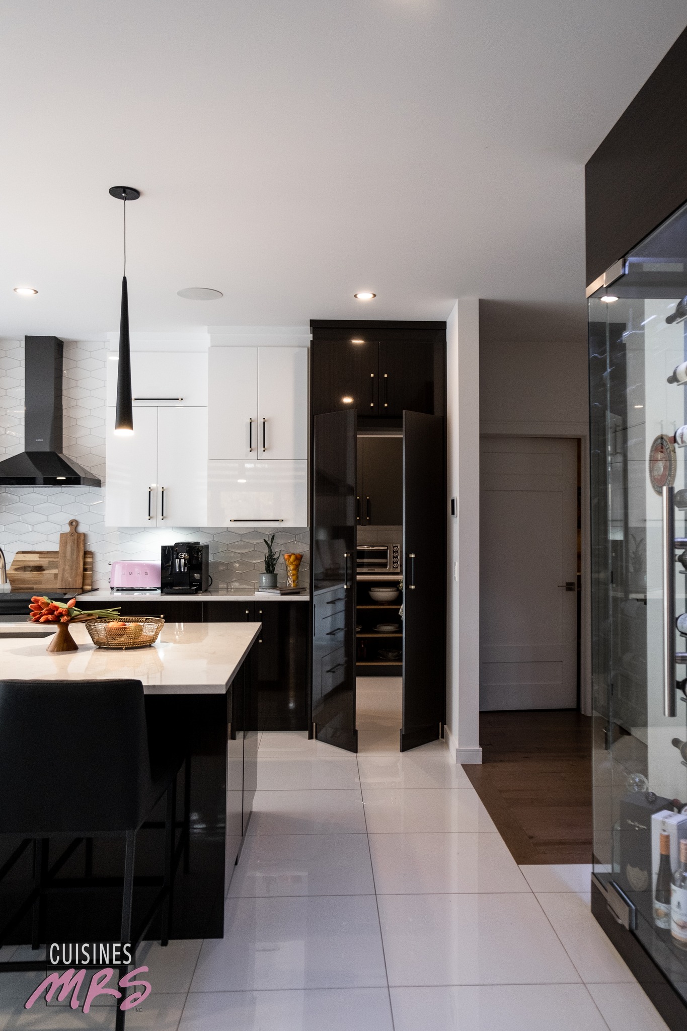 Cuisine moderne avec des armoires blanches et foncées, un îlot avec des chaises, des appareils électroménagers en acier inoxydable, des portes de garde-manger ouvertes et divers objets sur les comptoirs. Une lampe suspendue noire est accrochée au-dessus de l'îlot. Logo 