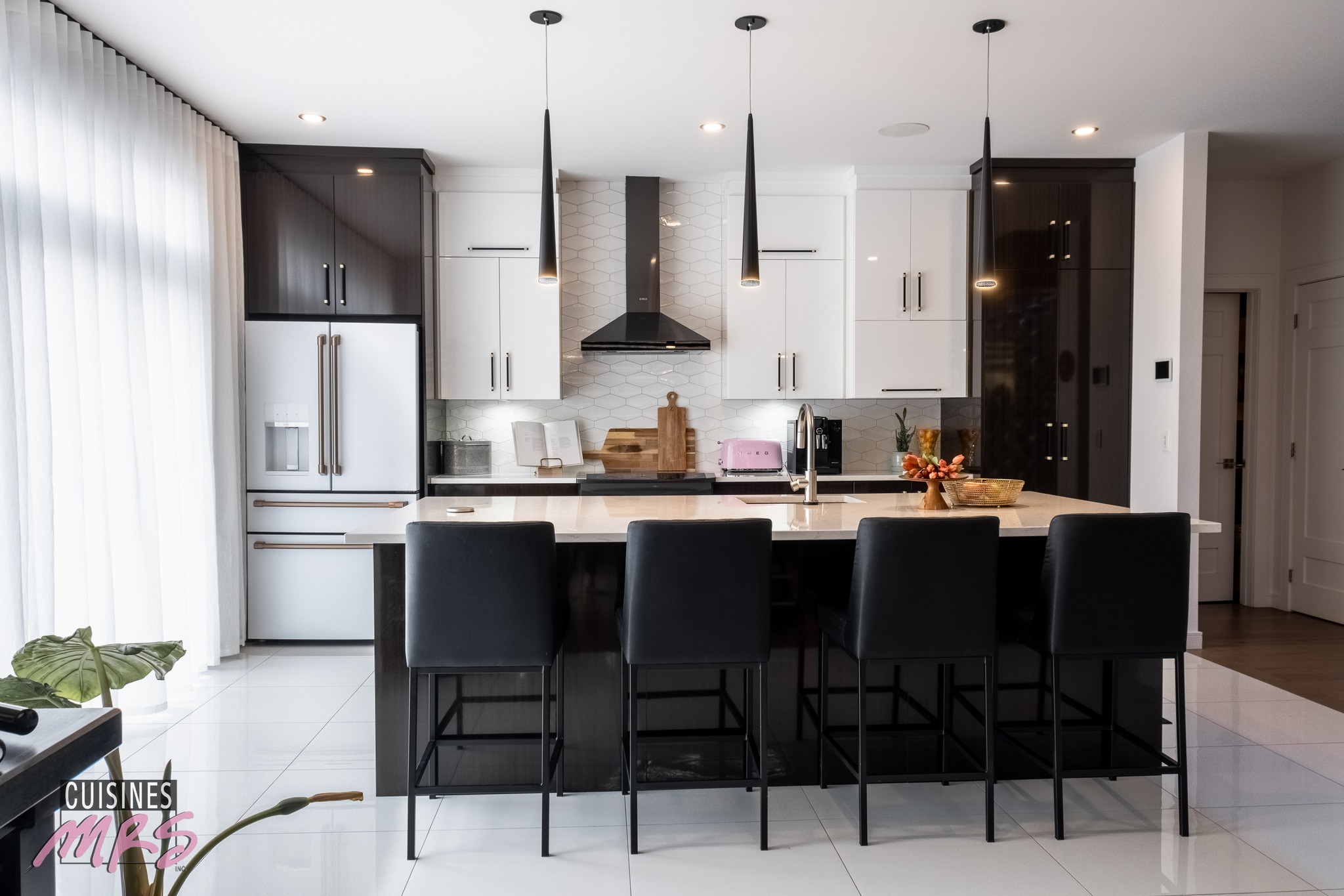Cuisine moderne avec des armoires noires et blanches, un îlot central avec quatre chaises noires, des lampes suspendues, un réfrigérateur, une cuisinière avec hotte, et des accessoires de comptoir comme un grille-pain rose et un bol à fruits. De grandes fenêtres apportent de la lumière naturelle.