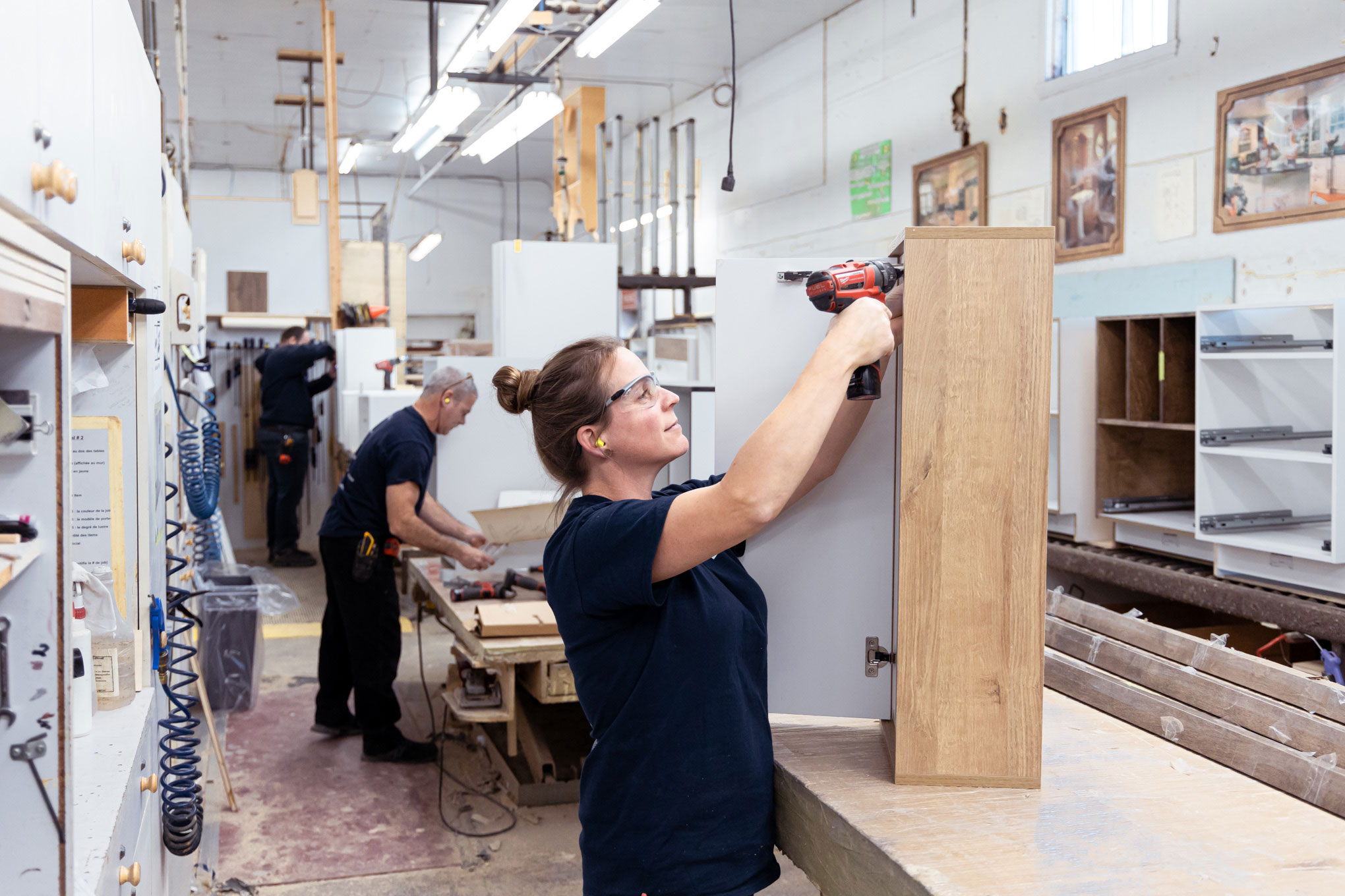 Une femme utilise une perceuse électrique sur une armoire en bois dans un atelier tandis qu'un homme travaille sur un autre projet à l'arrière-plan. L'atelier est rempli d'outils, d'armoires et de matériaux en bois.