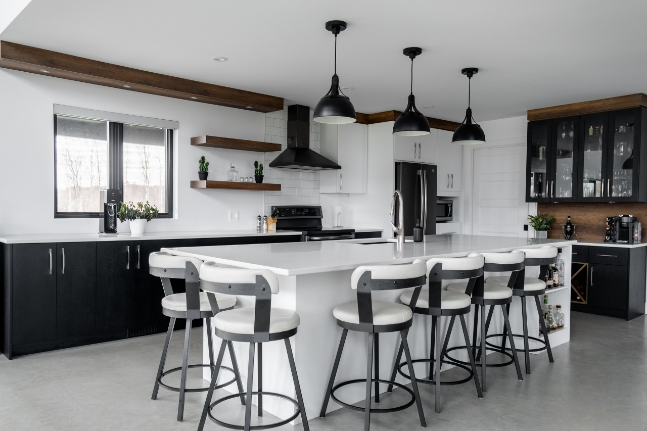 Cuisine moderne avec un grand îlot blanc, cinq tabourets de bar gris, des armoires noires et blanches, des appareils en acier inoxydable, des lampes suspendues, des étagères ouvertes avec des plantes et une fenêtre laissant entrer la lumière naturelle.