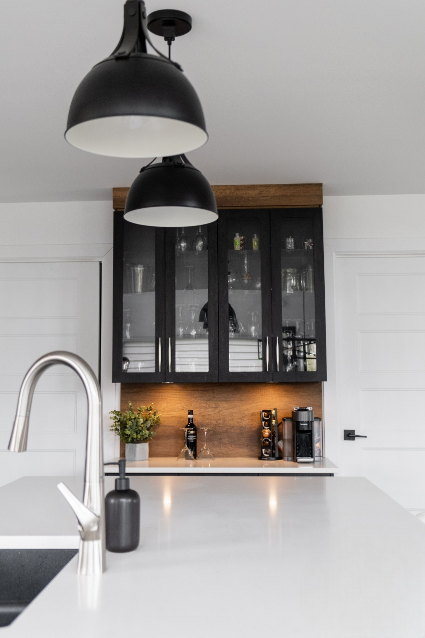 Cuisine moderne avec un plan de travail blanc, un évier et un robinet en acier inoxydable, des lampes suspendues noires et un meuble à portes vitrées présentant des verres et des bouteilles. Une machine à café et une plante se trouvent sur le comptoir arrière.