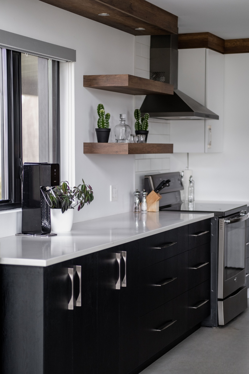 Cuisine moderne avec des armoires basses noires, un four en acier inoxydable, des comptoirs blancs, des étagères flottantes en bois contenant des bocaux et de petites plantes de cactus, une fenêtre et une plante en pot à côté d'un appareil électroménager noir sur le comptoir.
