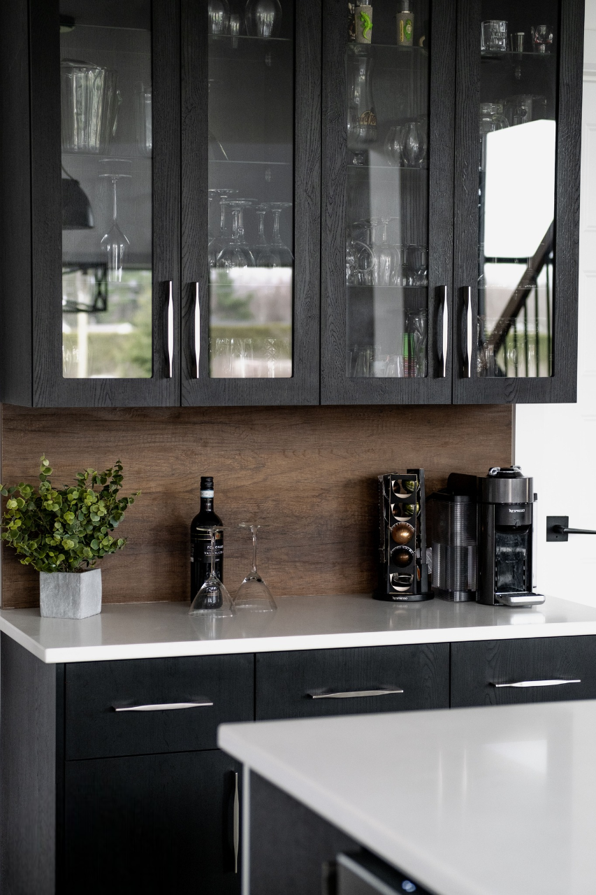 Un comptoir de cuisine moderne avec une plante en pot, une bouteille, deux verres à vin, un porte-capsule de café et une machine à café sous des armoires en verre foncé présentant de la verrerie.