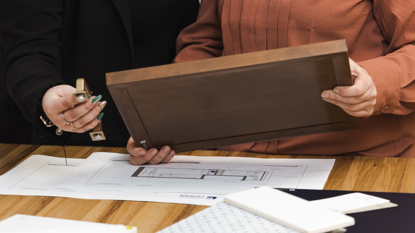 Deux personnes examinent un échantillon de porte d'armoire en bois et un ruban à mesurer sur des plans architecturaux posés sur une table en bois, afin d'évaluer les matériaux et les options de conception.