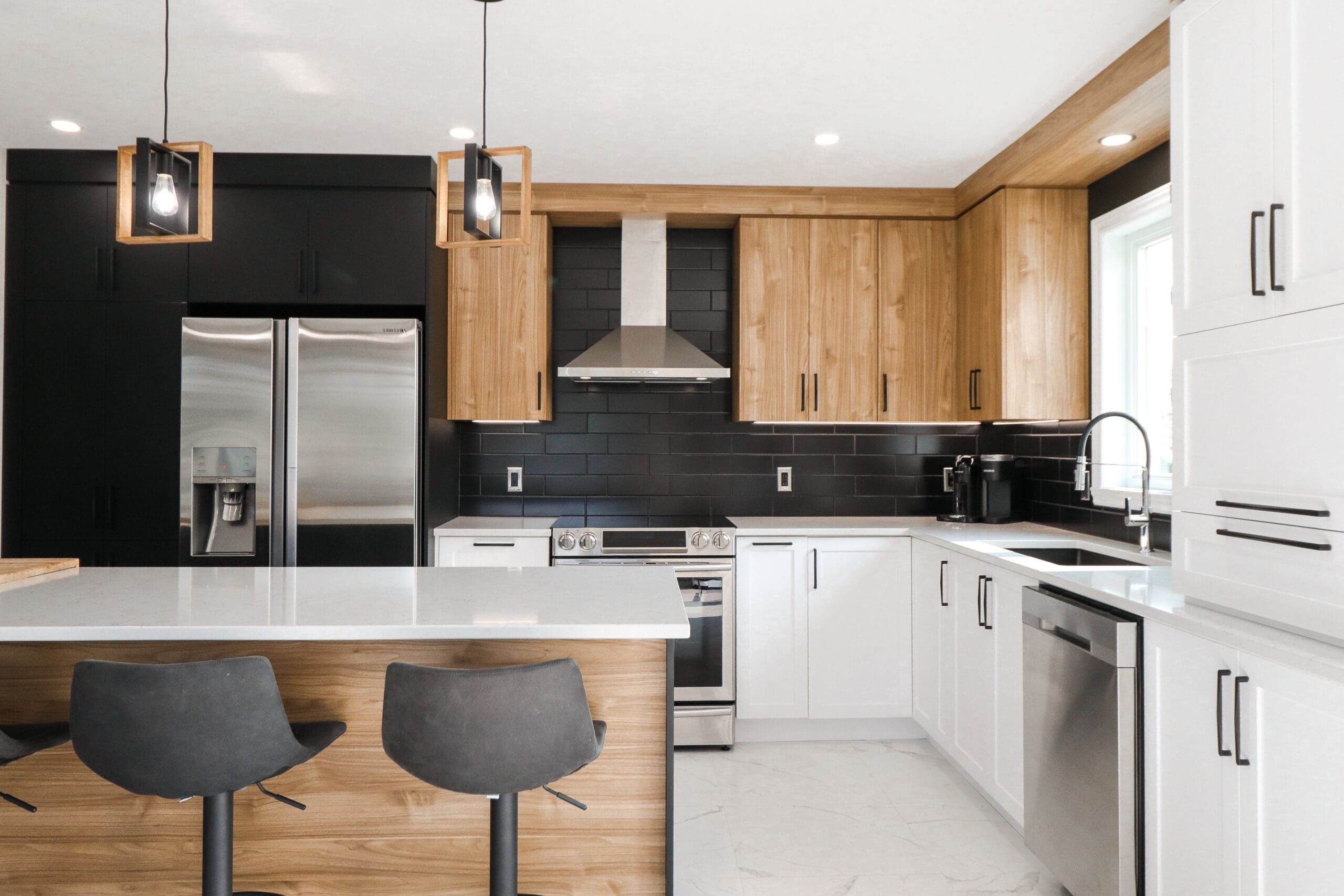 Cuisine moderne avec un mélange d'armoires en bois, noires et blanches, des appareils en acier inoxydable, un îlot central avec deux tabourets de bar sombres, des lampes suspendues et une fenêtre au-dessus de l'évier.