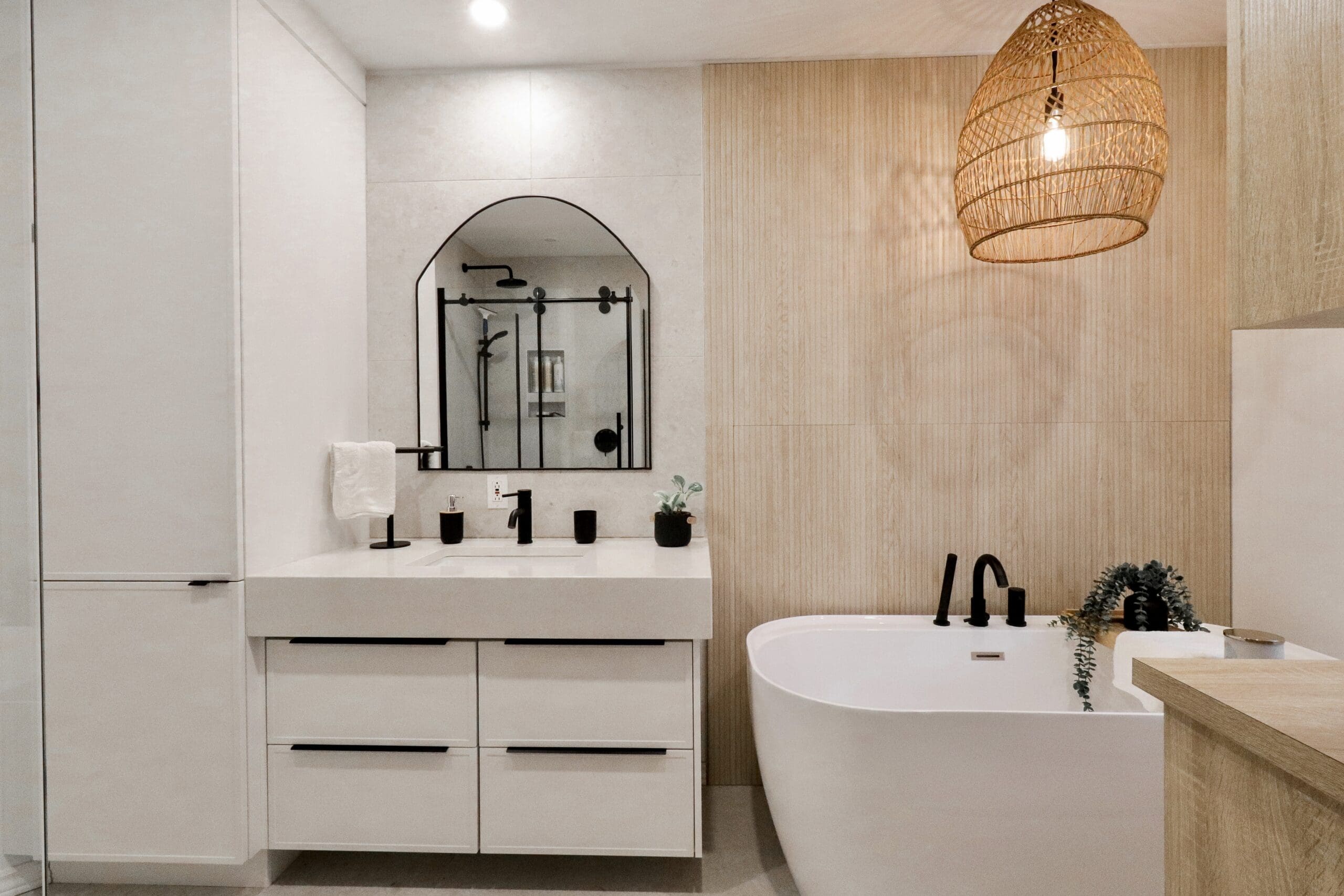 Salle de bains moderne avec un meuble-lavabo blanc, un miroir arqué, des luminaires noirs, une baignoire autoportante, une plante en pot, un mur d'accentuation en bois et une lampe suspendue tissée accrochée au plafond.