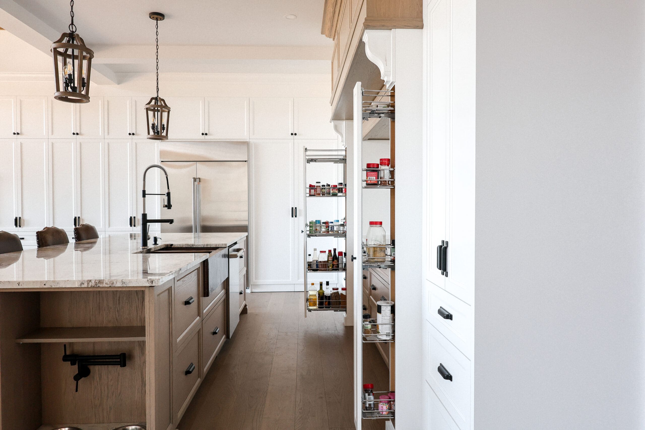 Une cuisine moderne comprend des armoires blanches, un grand îlot avec un évier intégré, des lampes suspendues et deux hautes étagères de garde-manger coulissantes remplies de divers produits alimentaires. La lumière naturelle éclaire cet espace spacieux et bien organisé.
