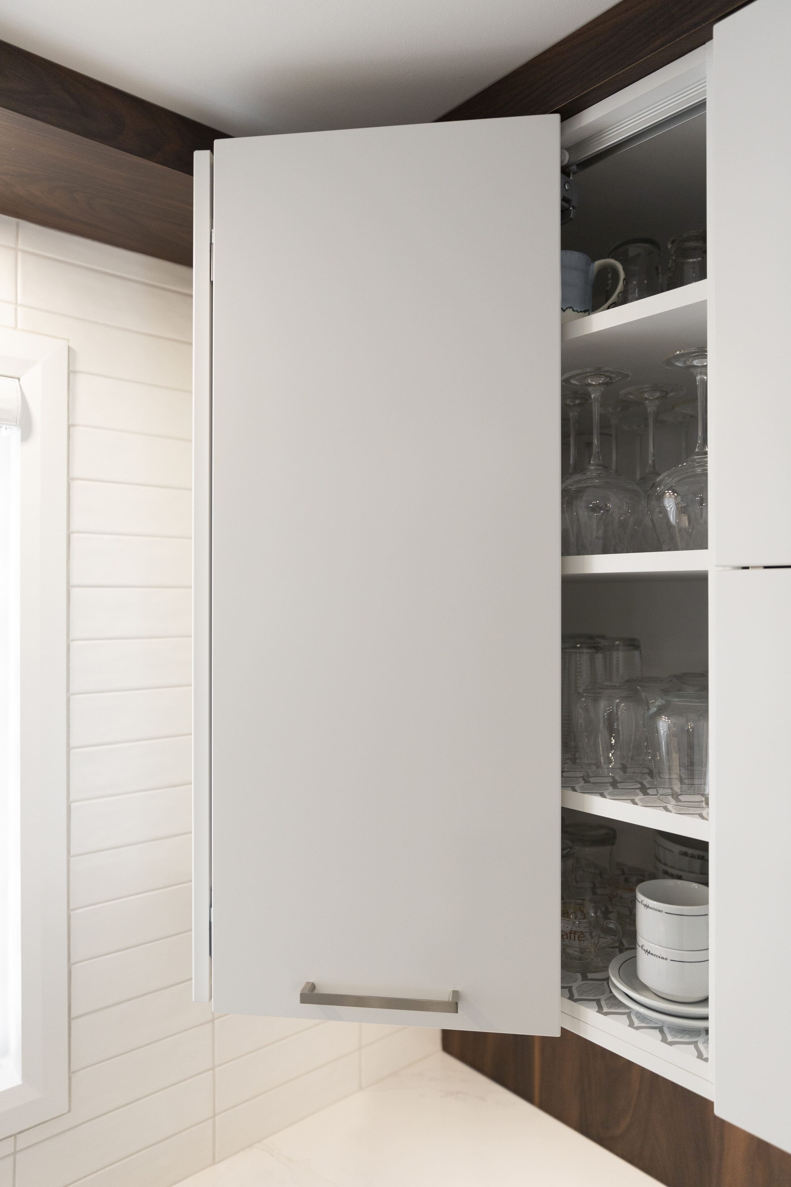 Un meuble de cuisine blanc dont la porte est partiellement ouverte, révélant des étagères contenant des verres, des tasses et des bols. Le meuble est monté sur un mur carrelé à côté d'une fenêtre.