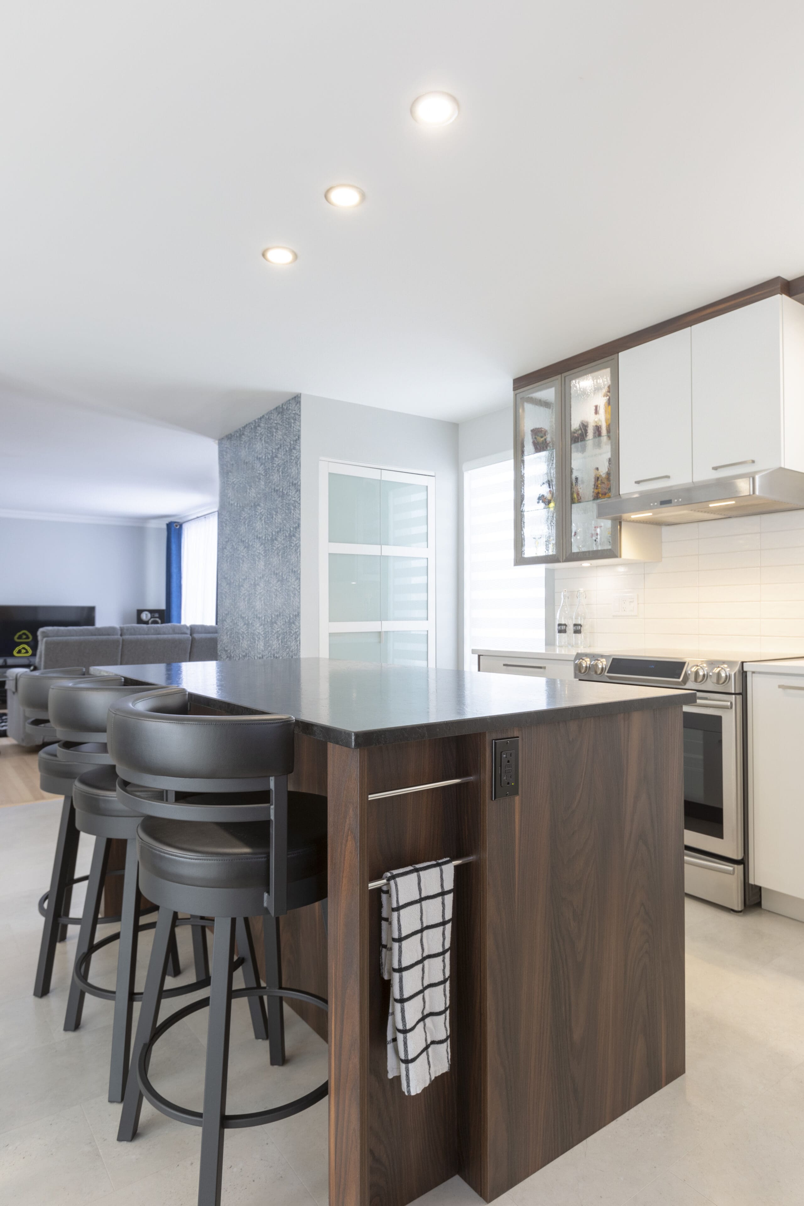 Cuisine moderne avec un îlot en bois foncé, quatre tabourets noirs, une serviette rayée suspendue sur le côté, des placards blancs, des appareils électroménagers en acier inoxydable et un éclairage zénithal lumineux.