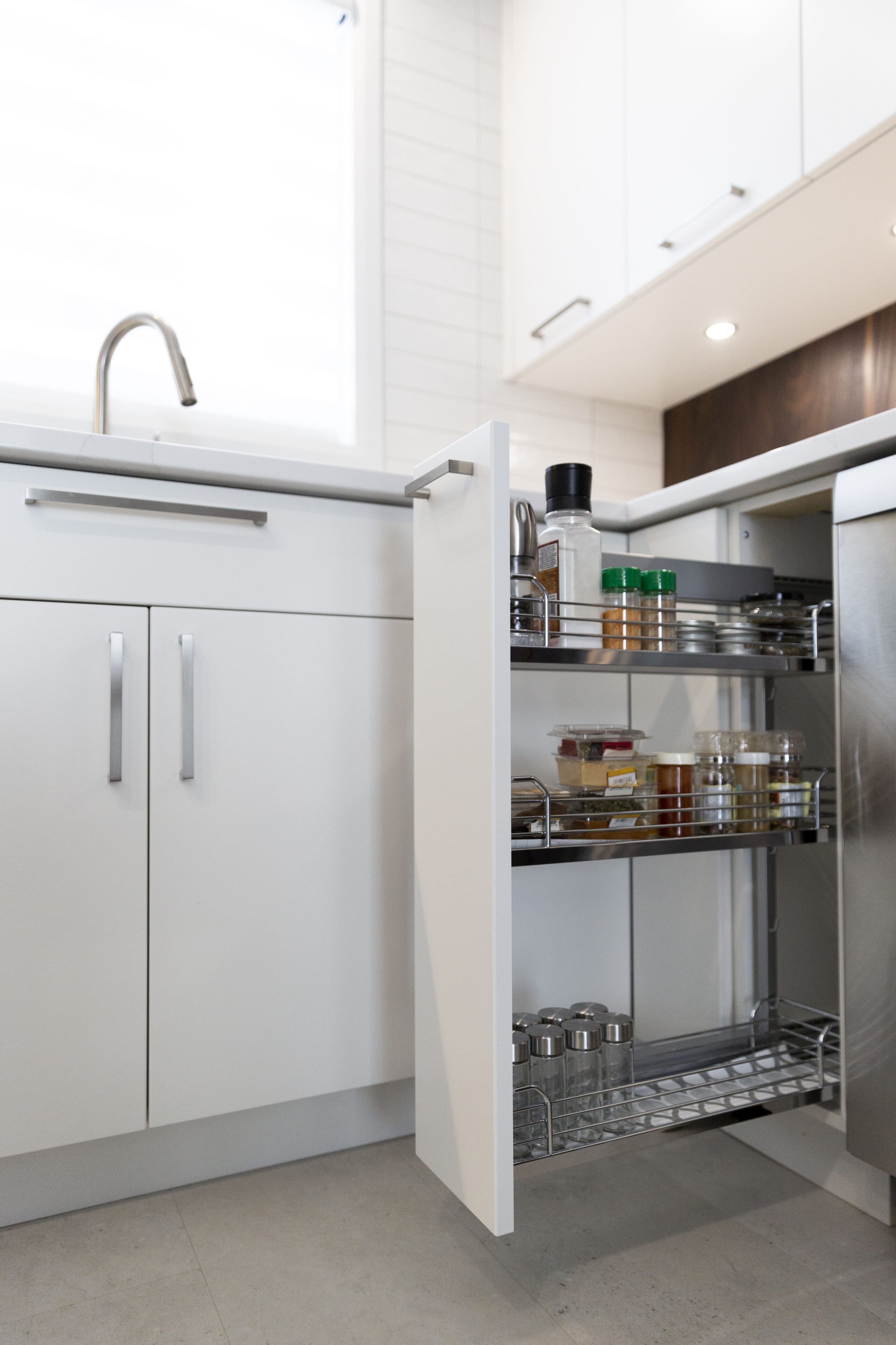 Une cuisine blanche moderne avec une armoire à tiroirs ouverte, présentant des épices et des condiments bien rangés sur des étagères en métal. Le plan de travail, l'évier et les armoires sont propres et minimalistes.
