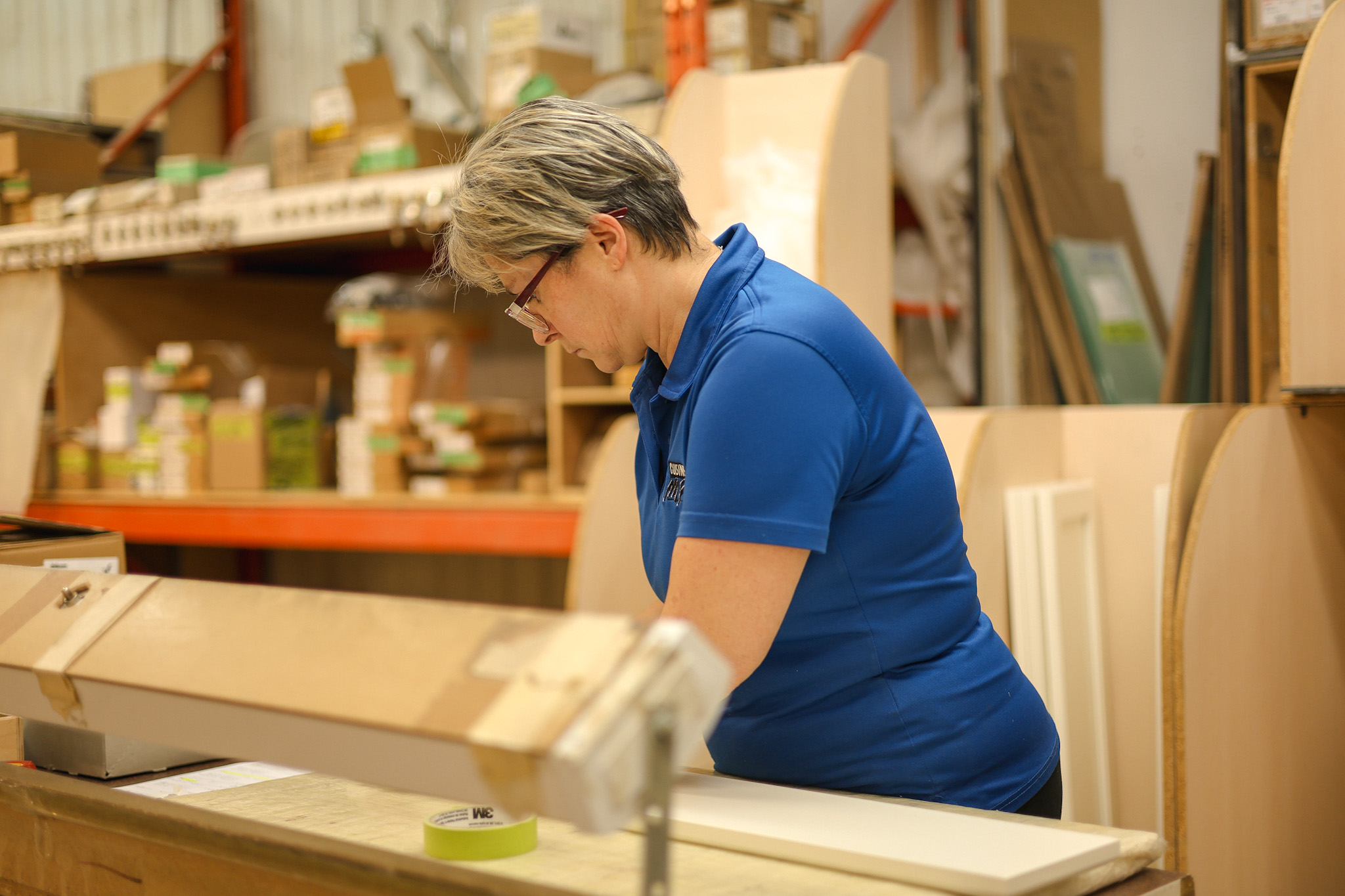 Une personne aux cheveux courts et aux lunettes, portant un polo bleu, travaille attentivement à une table dans un entrepôt ou un atelier rempli de boîtes, d'étagères et de divers matériaux de construction.