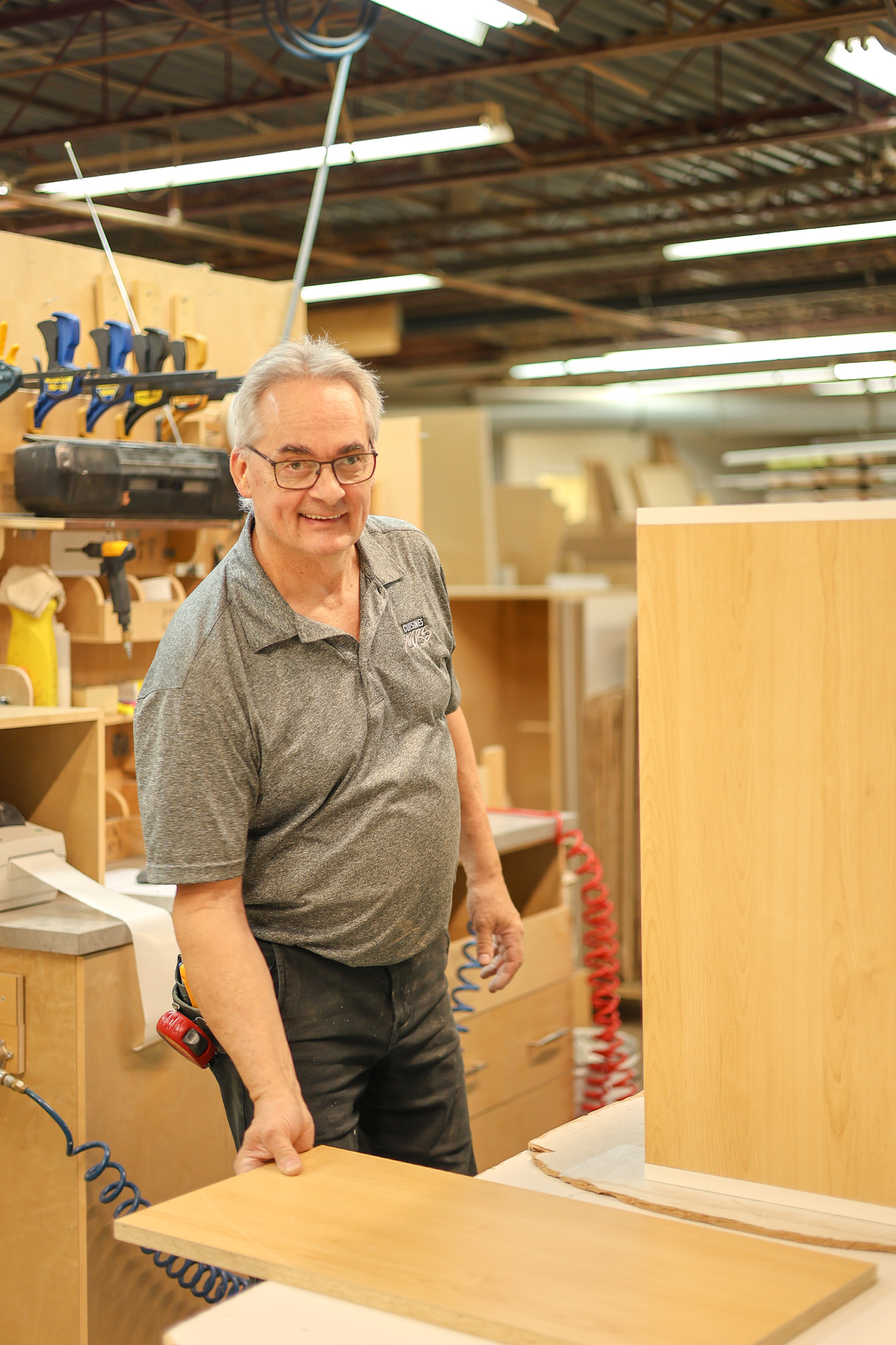 Un homme âgé portant des lunettes et un polo gris sourit en travaillant avec des planches de bois dans un atelier de menuiserie rempli d'outils et d'armoires.