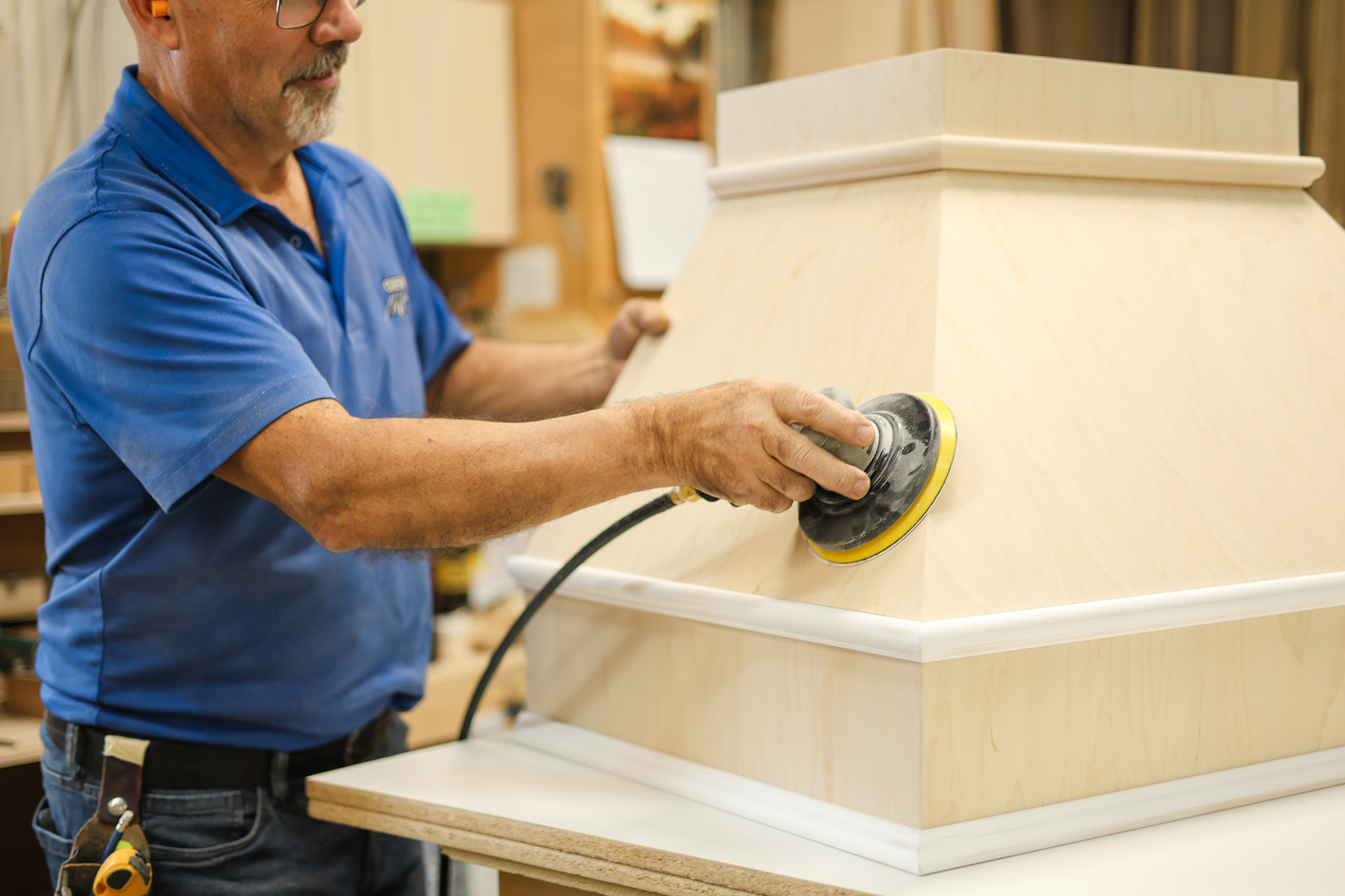 Un homme en chemise bleue utilise une ponceuse électrique pour lisser la surface d'une grande structure en bois clair dans un atelier.