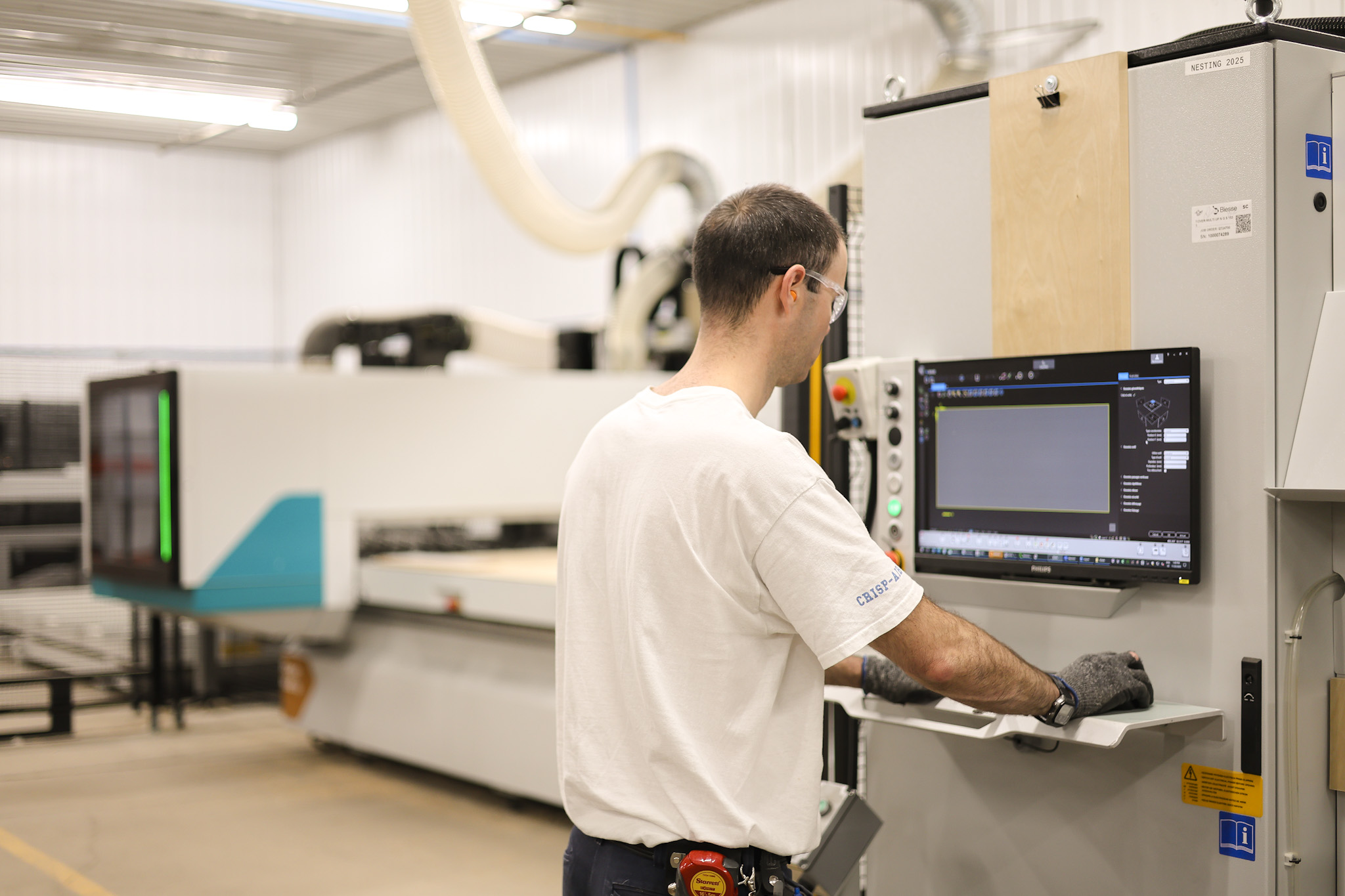 Un homme portant des gants et des lunettes de sécurité manipule un panneau de commande dans une installation industrielle moderne avec de grandes machines à l'arrière-plan.
