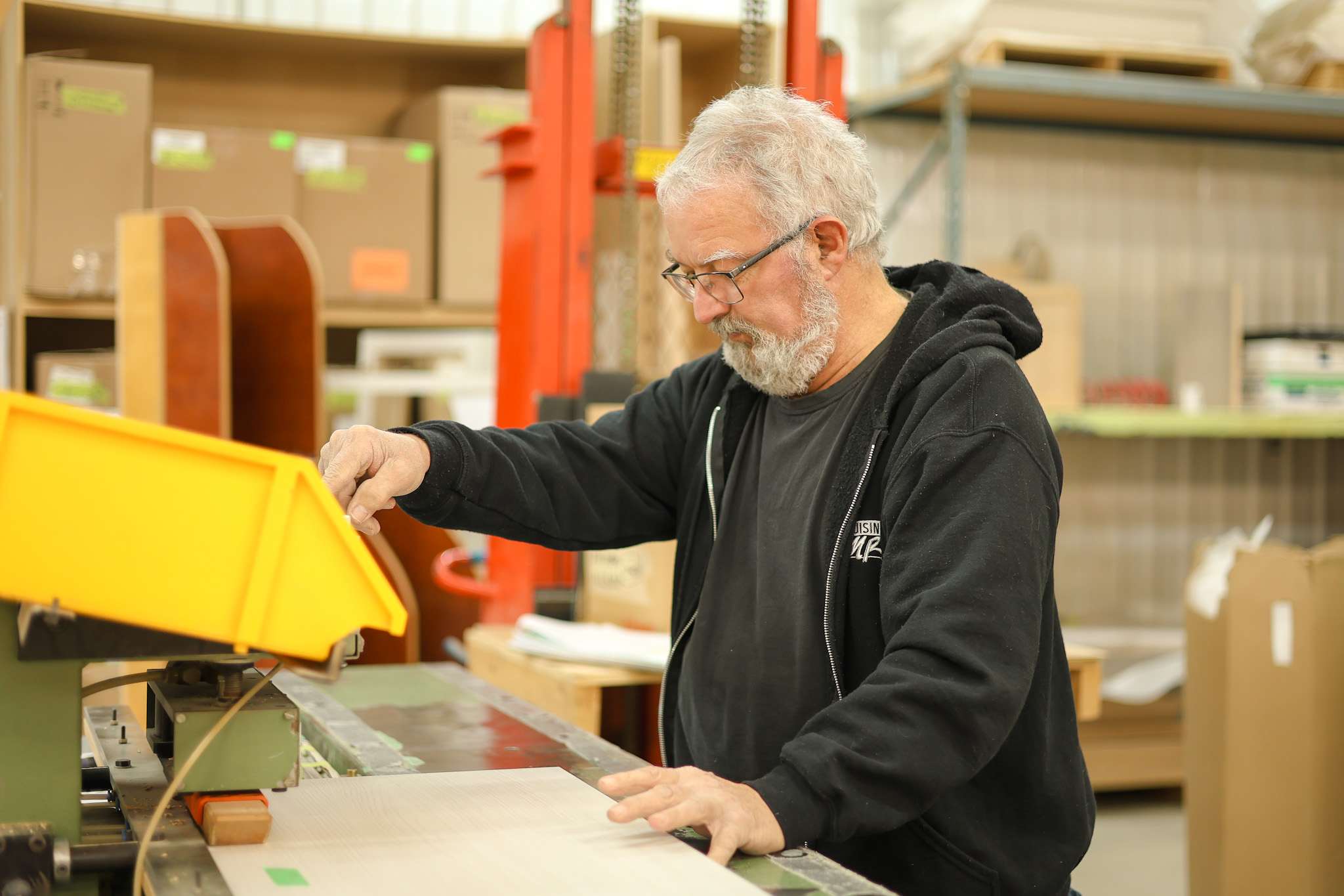 Dans un entrepôt, un homme âgé aux cheveux gris et aux lunettes actionne une machine, vêtu d'un sweat à capuche noir. Des étagères contenant des boîtes et des fournitures se trouvent à l'arrière-plan.