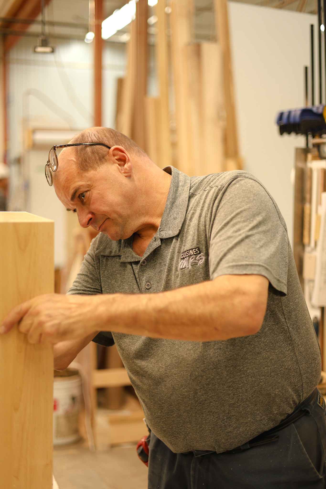 Un homme portant des lunettes et un polo gris travaille avec attention sur une planche de bois dans un atelier rempli d'outils et de matériaux de menuiserie.