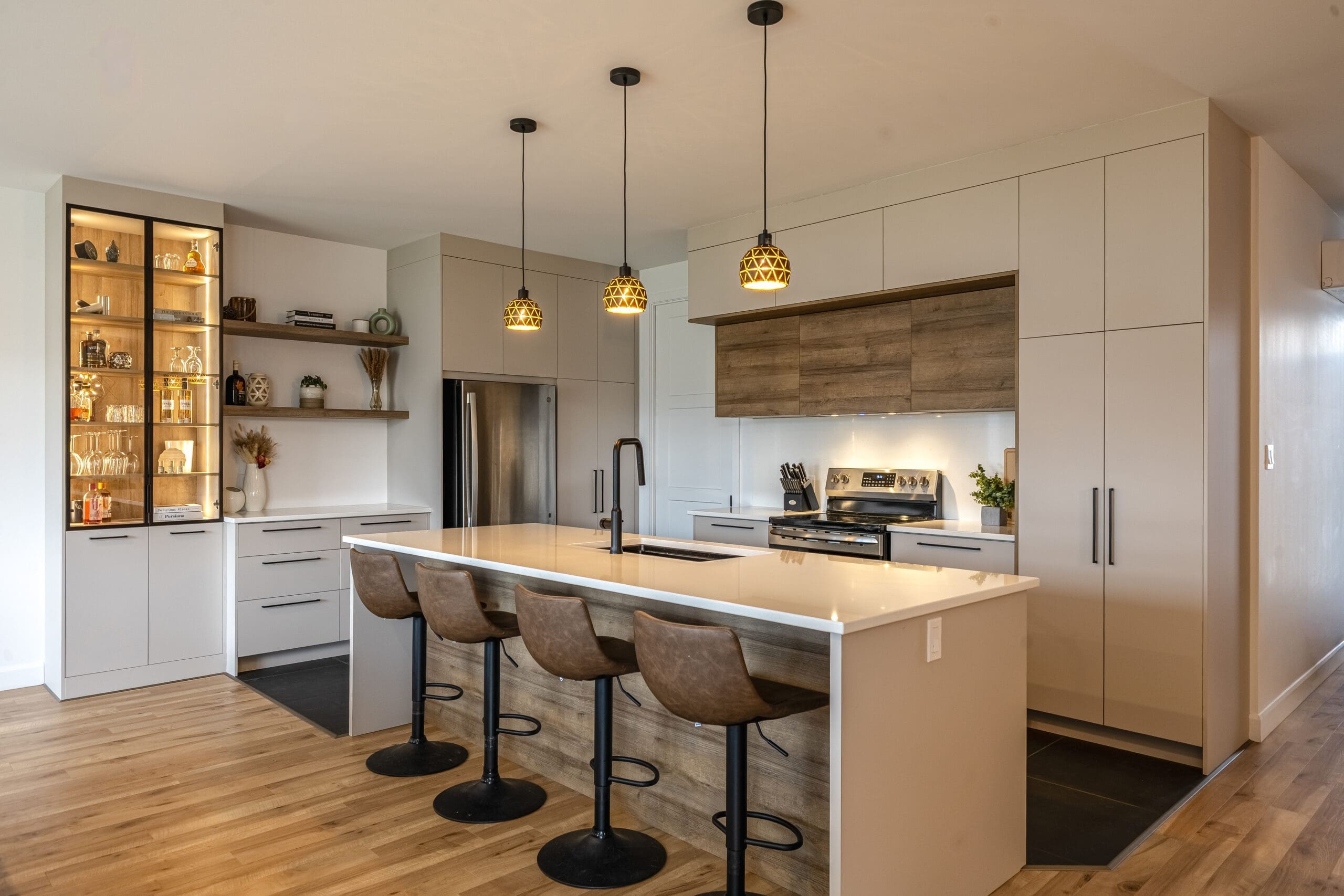 Cuisine moderne avec des armoires beige couleur sahara et melamine, un grand îlot avec quatre tabourets bruns, des lampes suspendues, des appareils électroménagers en acier inoxydable et des étagères ouvertes présentant des objets de décoration et des ustensiles de cuisine.