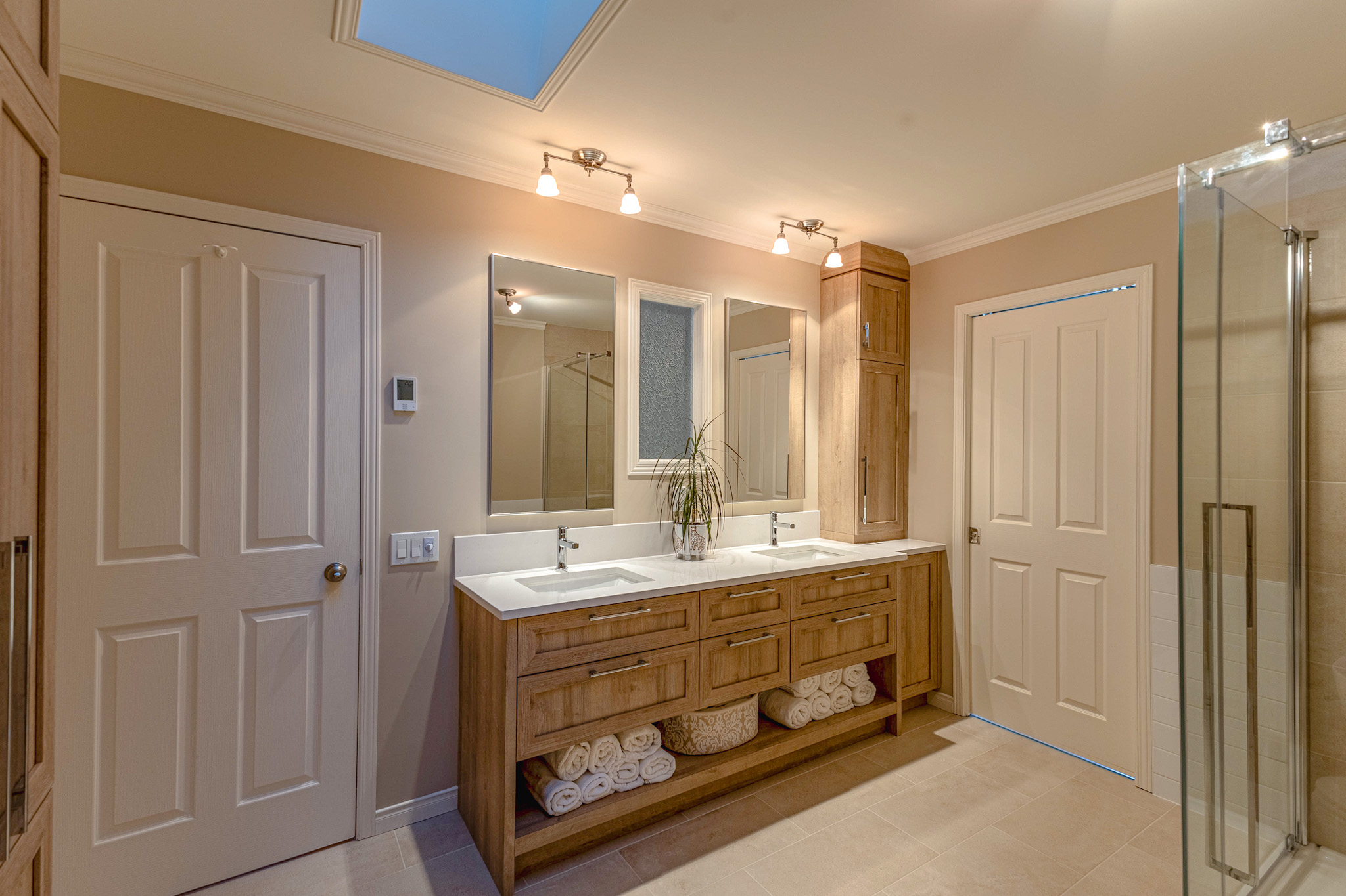 Salle de bains spacieuse avec meuble à double vasque, armoires en bois, serviettes enroulées sur des étagères, cabine de douche en verre et éclairage zénithal doux. Une lucarne et deux miroirs ajoutent de la luminosité à cette pièce aux tons neutres.