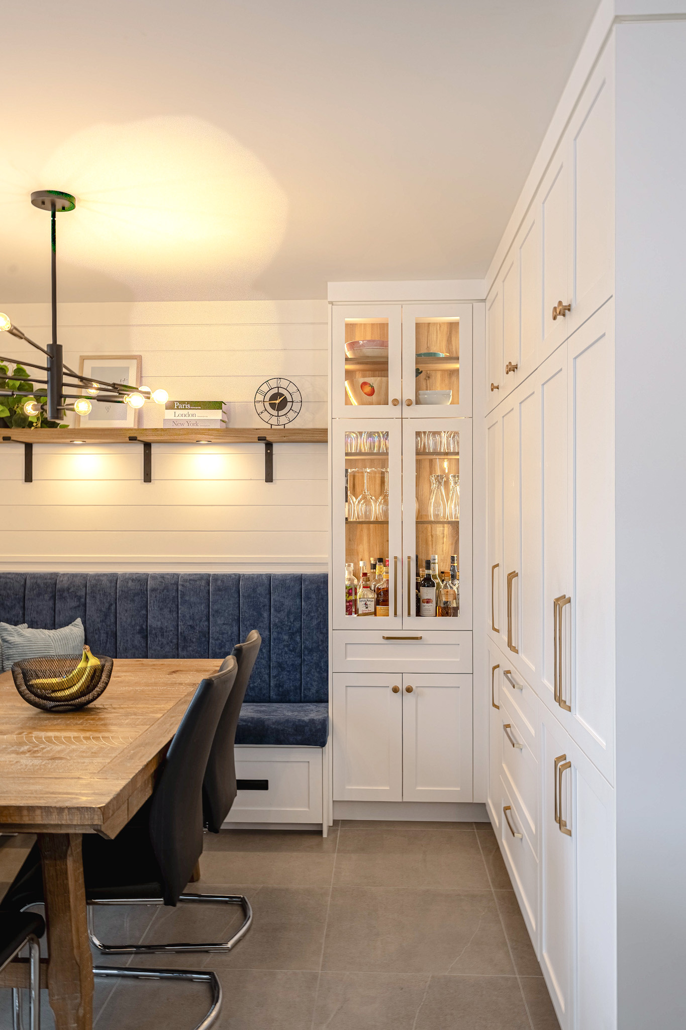 Une cuisine moderne avec des armoires blanches, une vitrine encastrée avec des boissons et des verres, une table à manger en bois, des chaises noires et une banquette bleue le long du mur. Un luminaire contemporain est suspendu au-dessus.