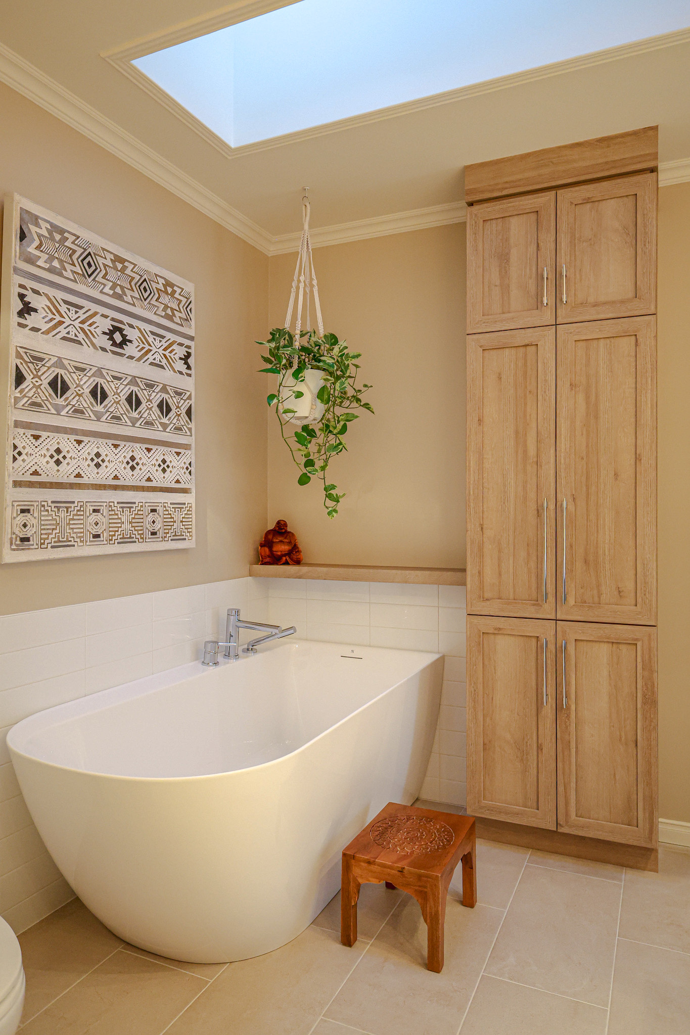 Salle de bains moderne avec une baignoire blanche autoportante, un meuble en bois, un tableau mural à motifs, une plante suspendue, un petit tabouret en bois et une lucarne qui apporte de la lumière naturelle.