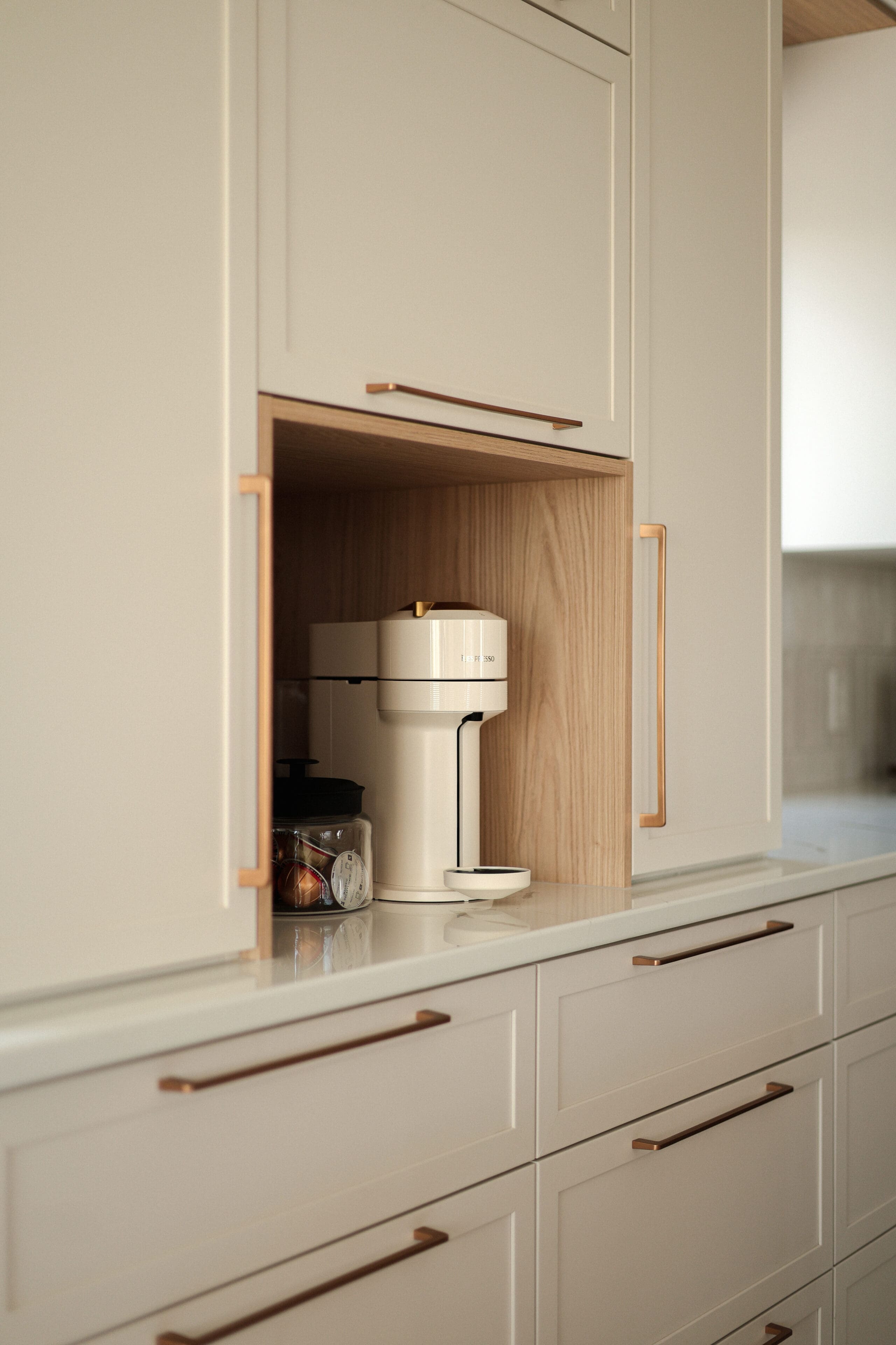 Un coin cuisine moderne avec une cafetière encastrée, un pot en verre contenant des dosettes de café et des armoires de couleur claire avec des poignées en cuivre élégantes.