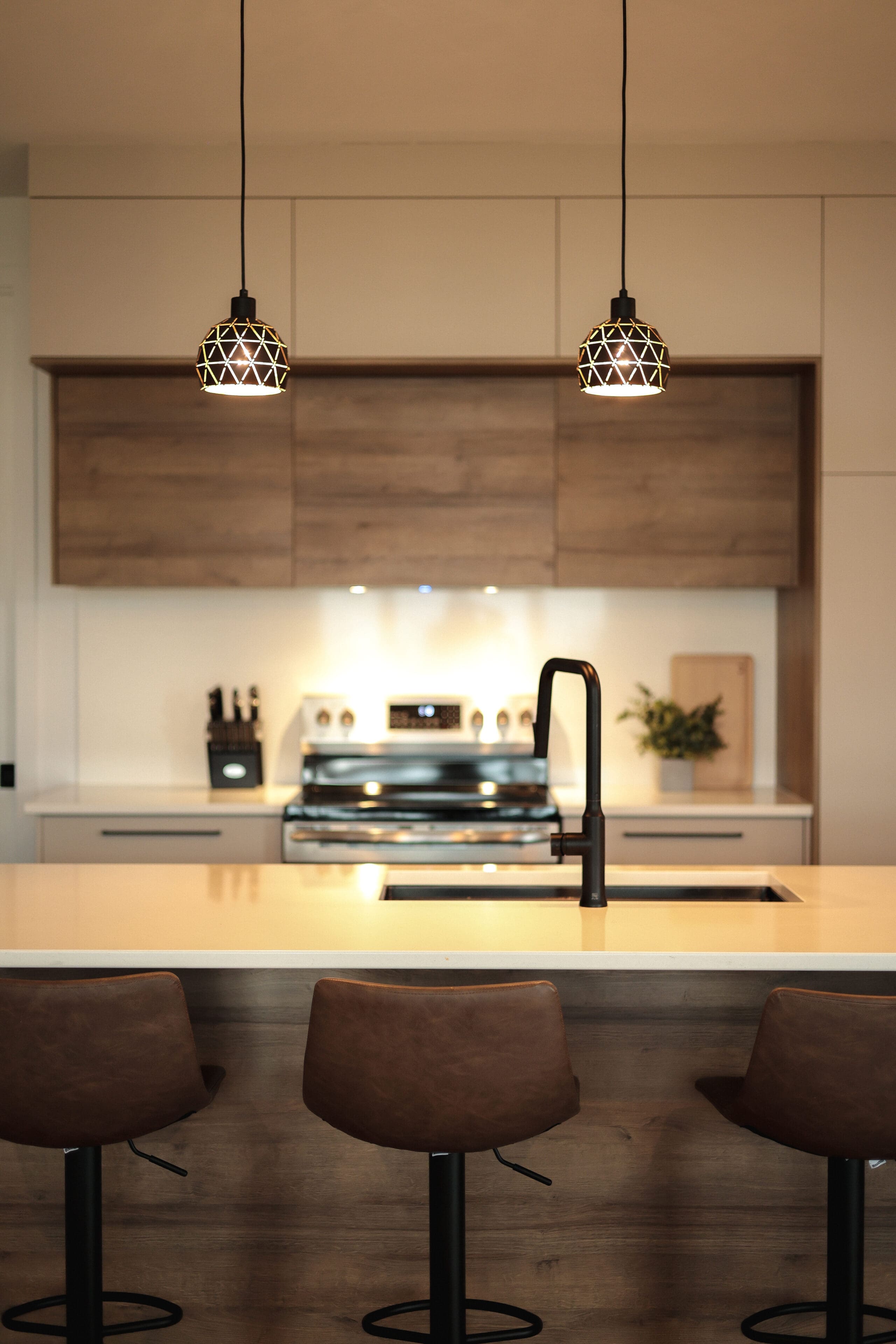 Cuisine moderne avec des armoires en bois, un îlot central avec trois tabourets de bar marron, un robinet noir, des appareils en acier inoxydable et deux lampes suspendues géométriques suspendues au-dessus du comptoir.